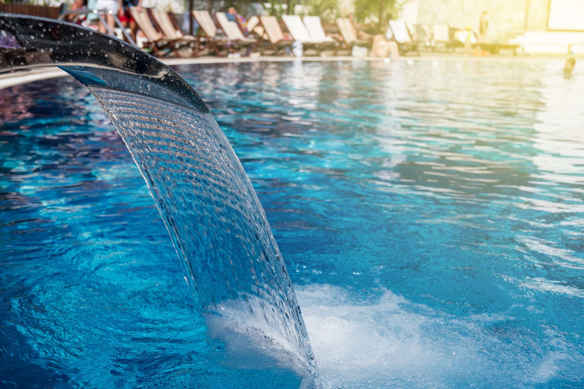 A chrome water fountain pours a sheet of water into a vibrant blue swimming pool with lounge chairs on the deck.