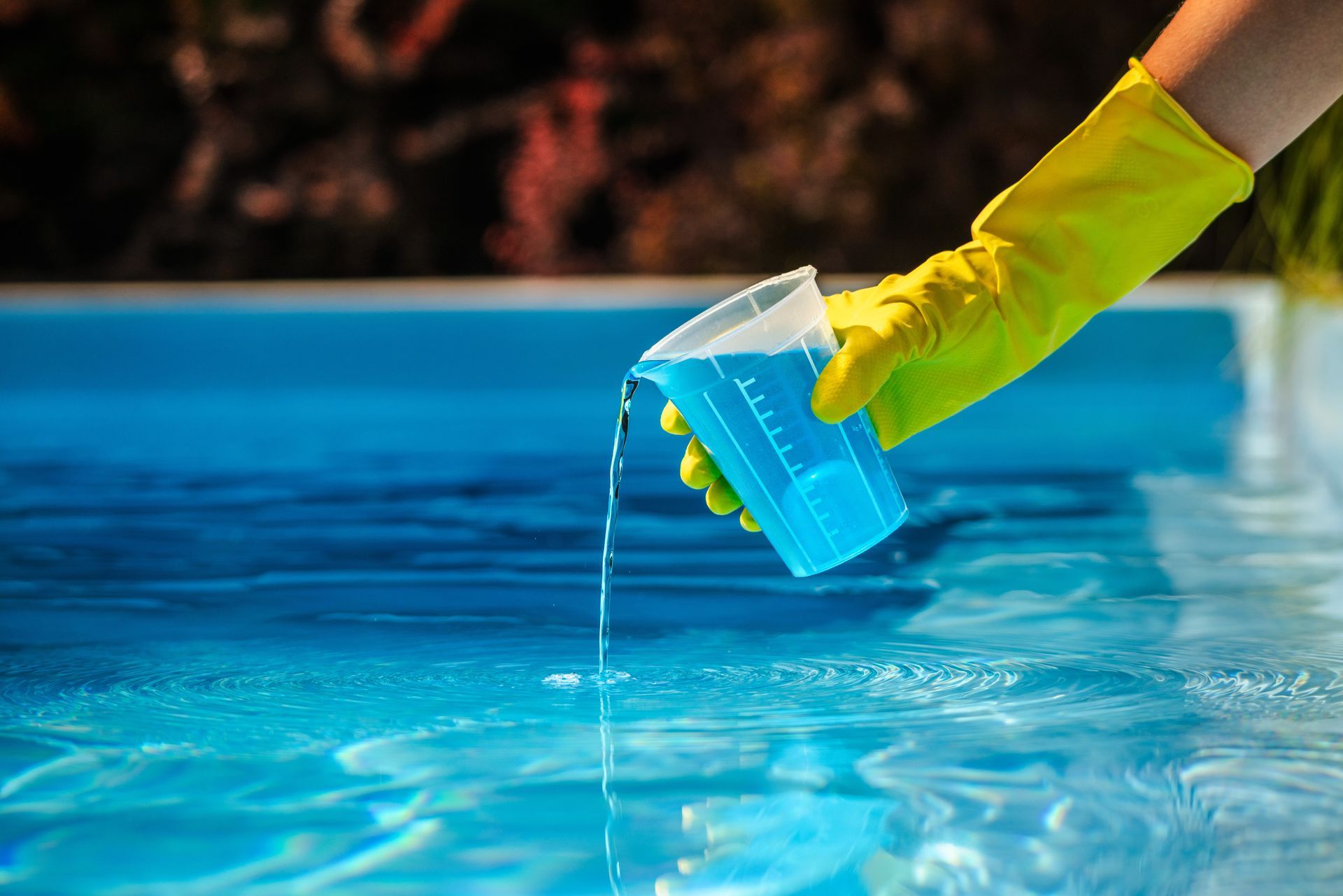 A person wearing a yellow rubber glove pours blue liquid from a measuring cup into a swimming pool.