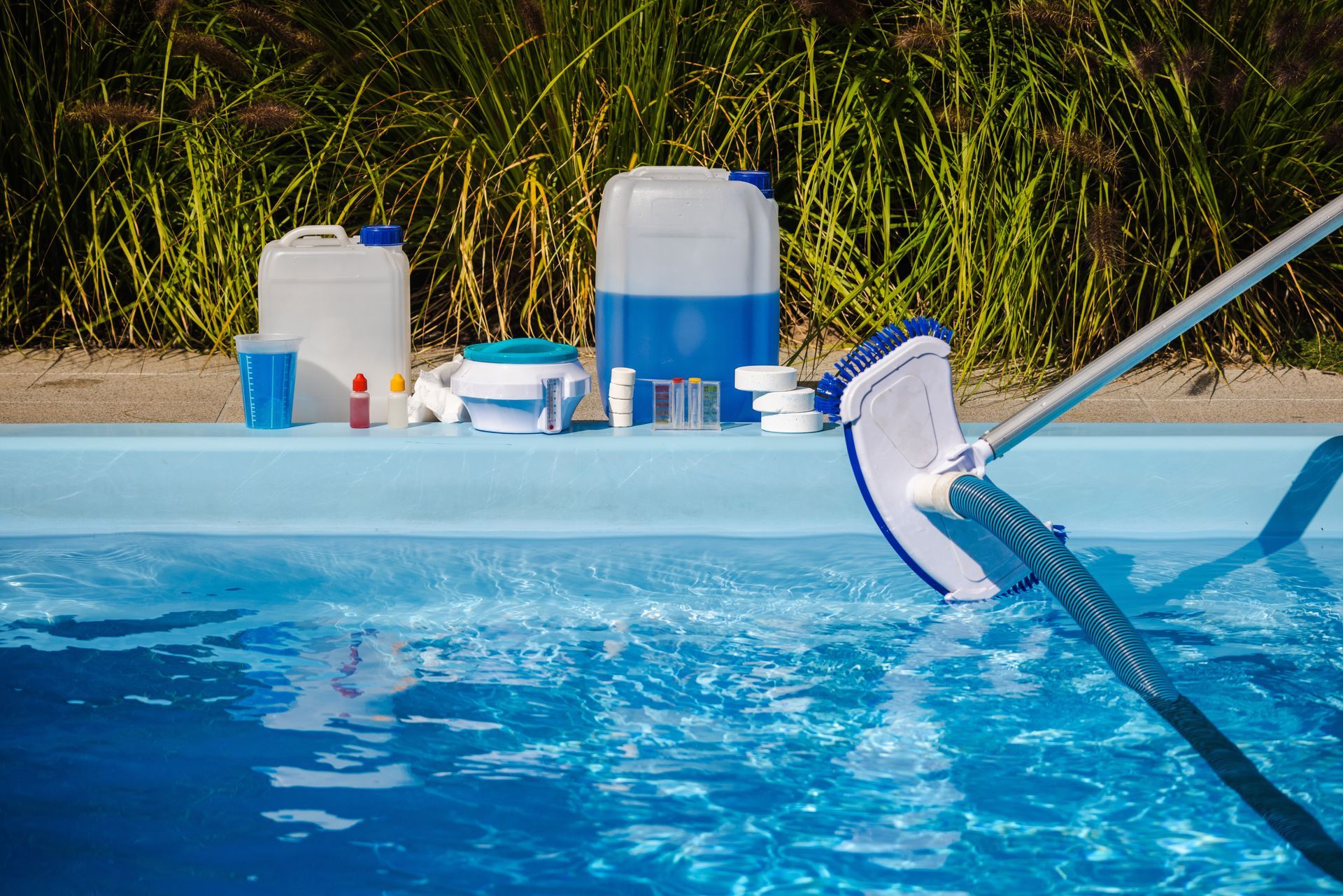 A pool brush rests in clear blue water, with maintenance chemicals and equipment arranged on the deck in the background.