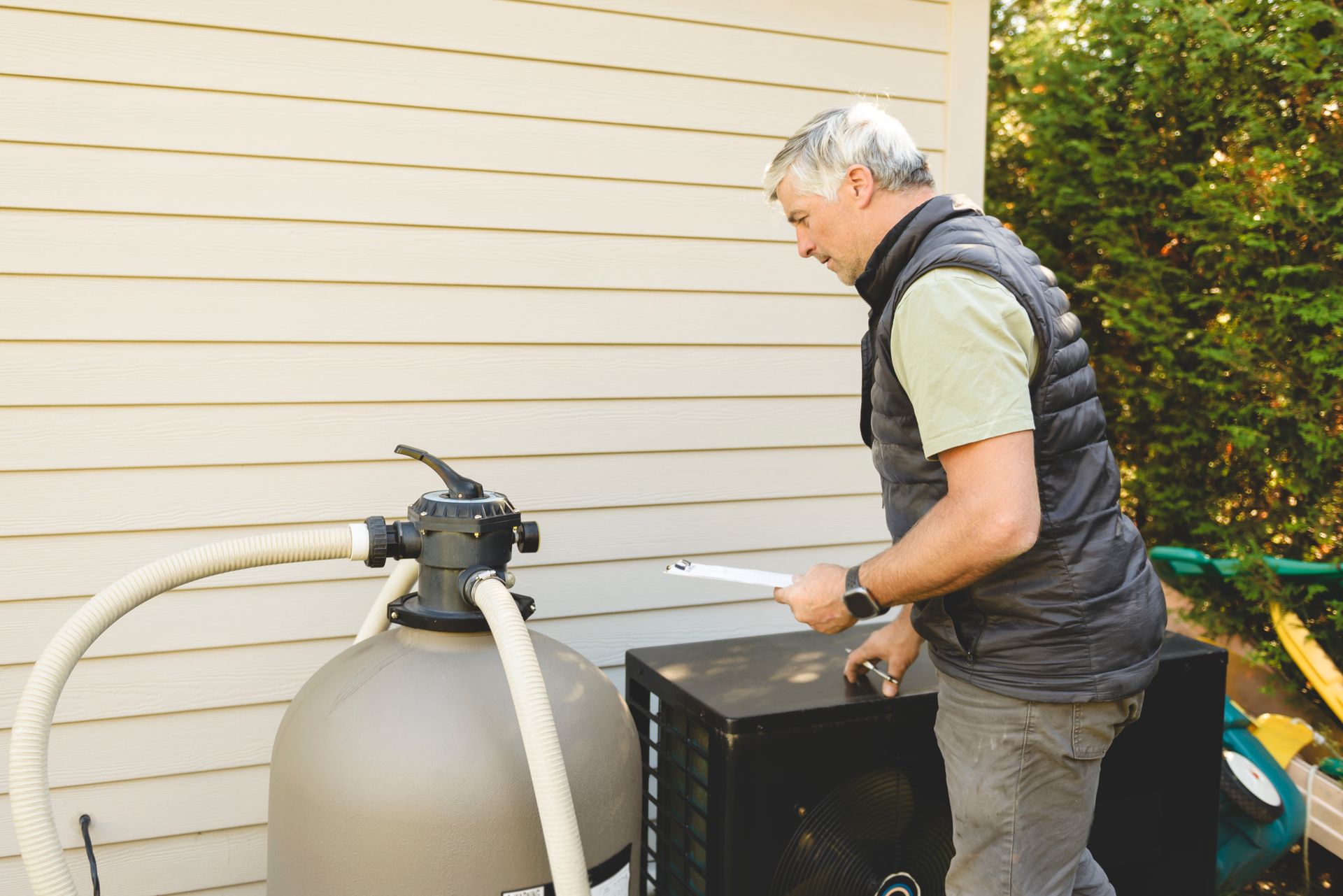 A person standing in a backyard, checking a pool pump and filter system next to a beige-sided house.