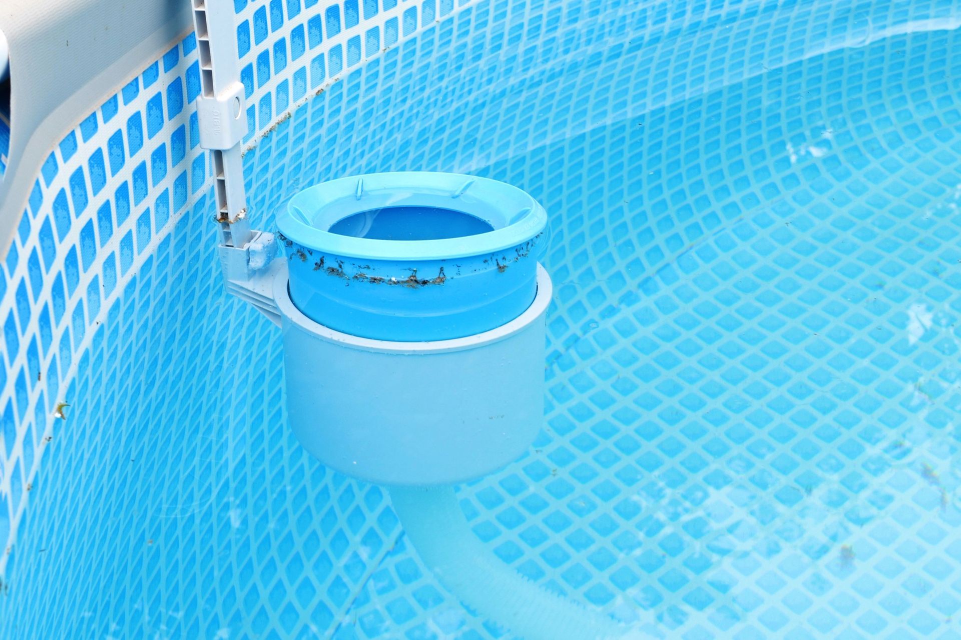 A blue and grey pool surface skimmer attached to the wall of a blue, tiled-patterned above-ground swimming pool.