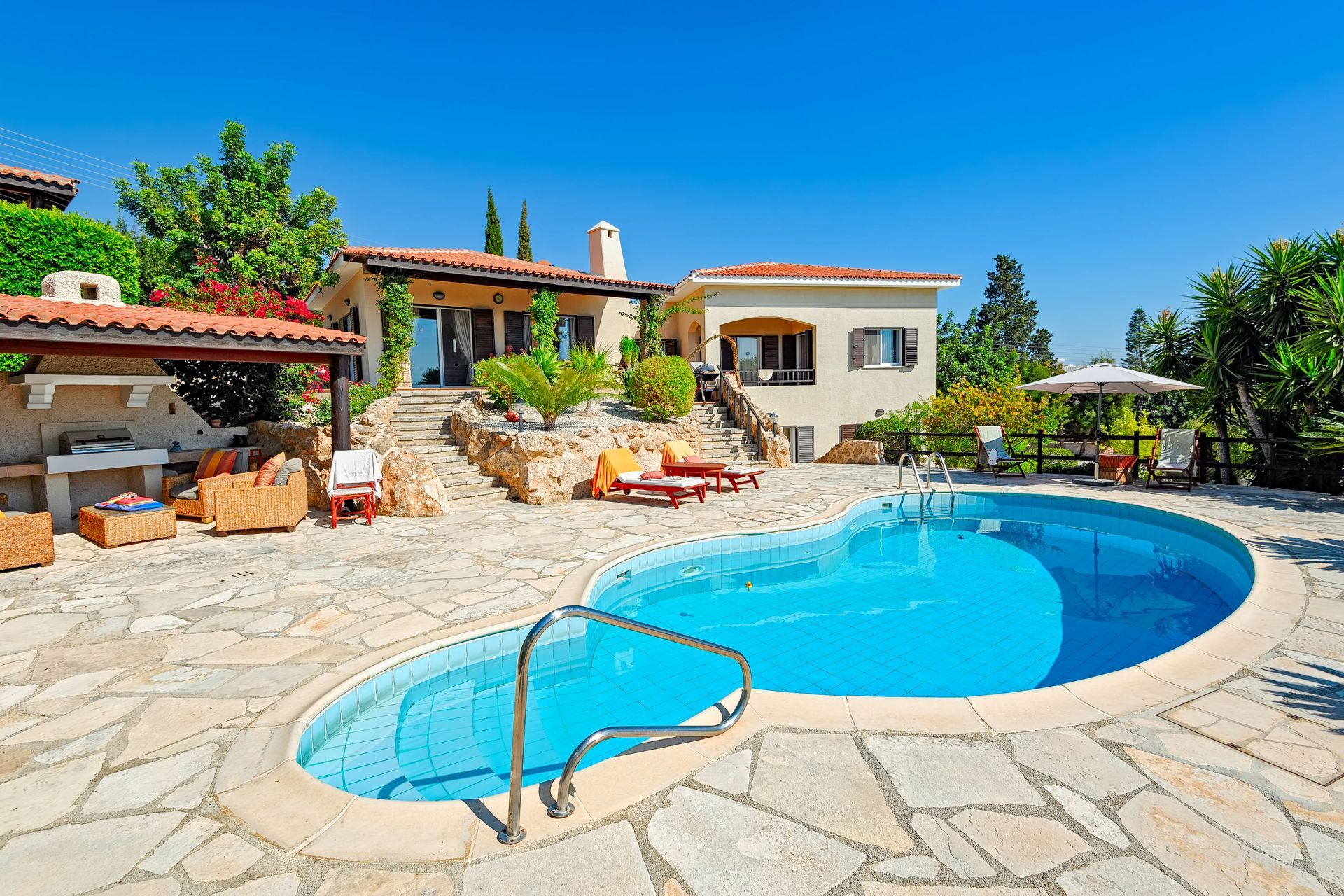 Sunny backyard with a blue swimming pool, stone patio, and Mediterranean-style house under a clear sky