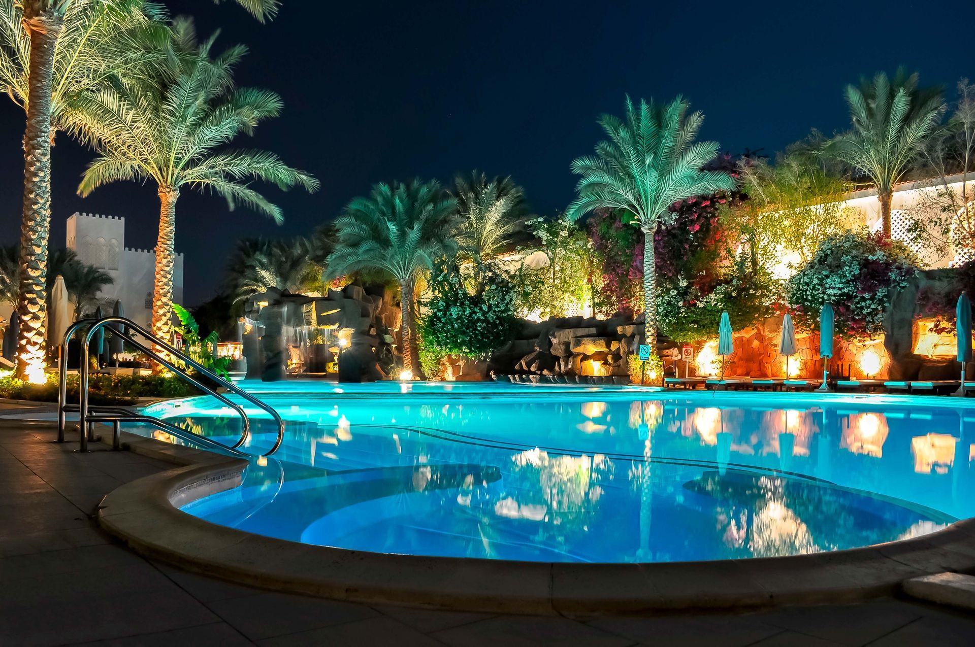 Illuminated tropical resort pool at night with palm trees and blue reflections