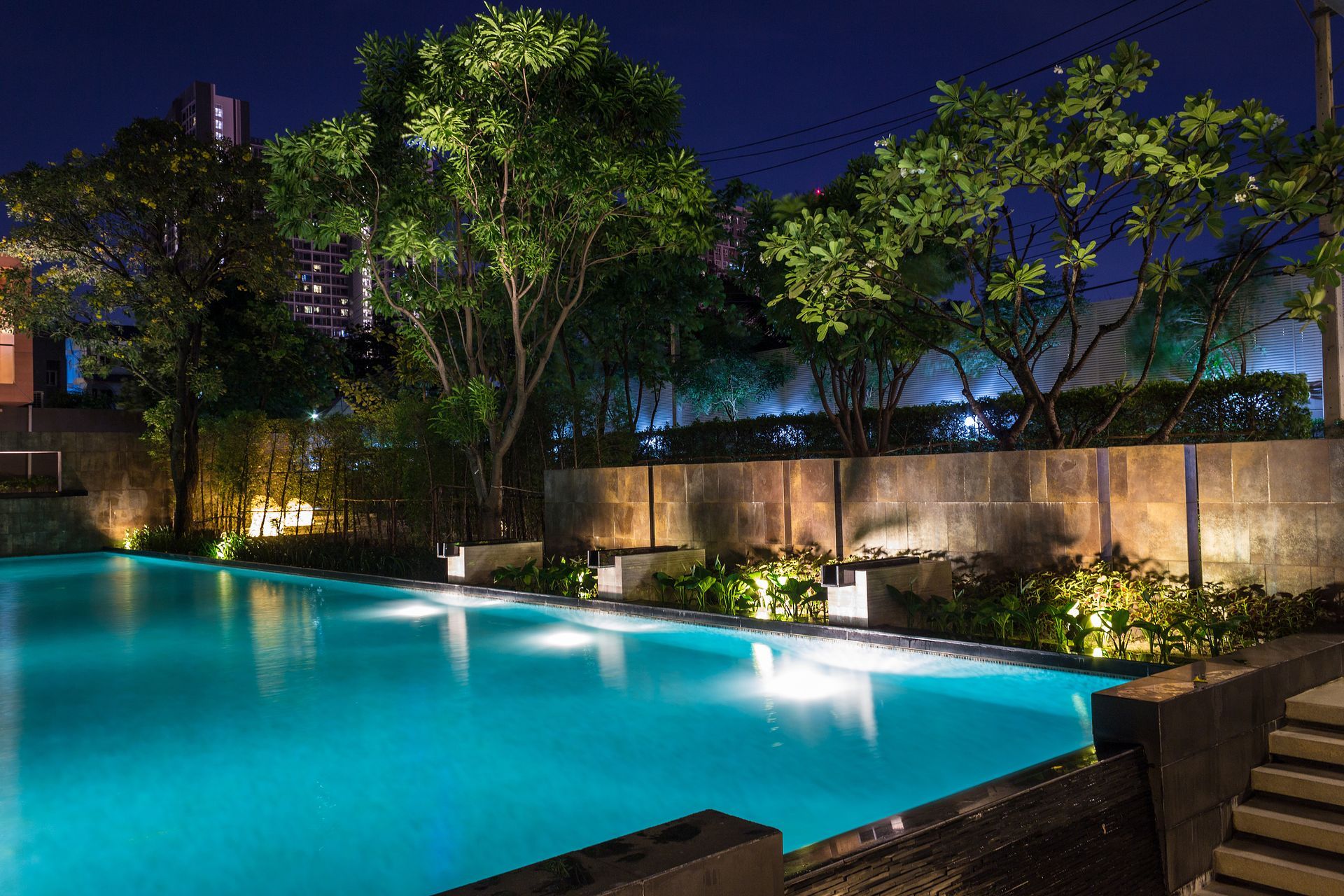 A brightly lit turquoise swimming pool at night, surrounded by lush trees and a stone wall with warm garden lighting.