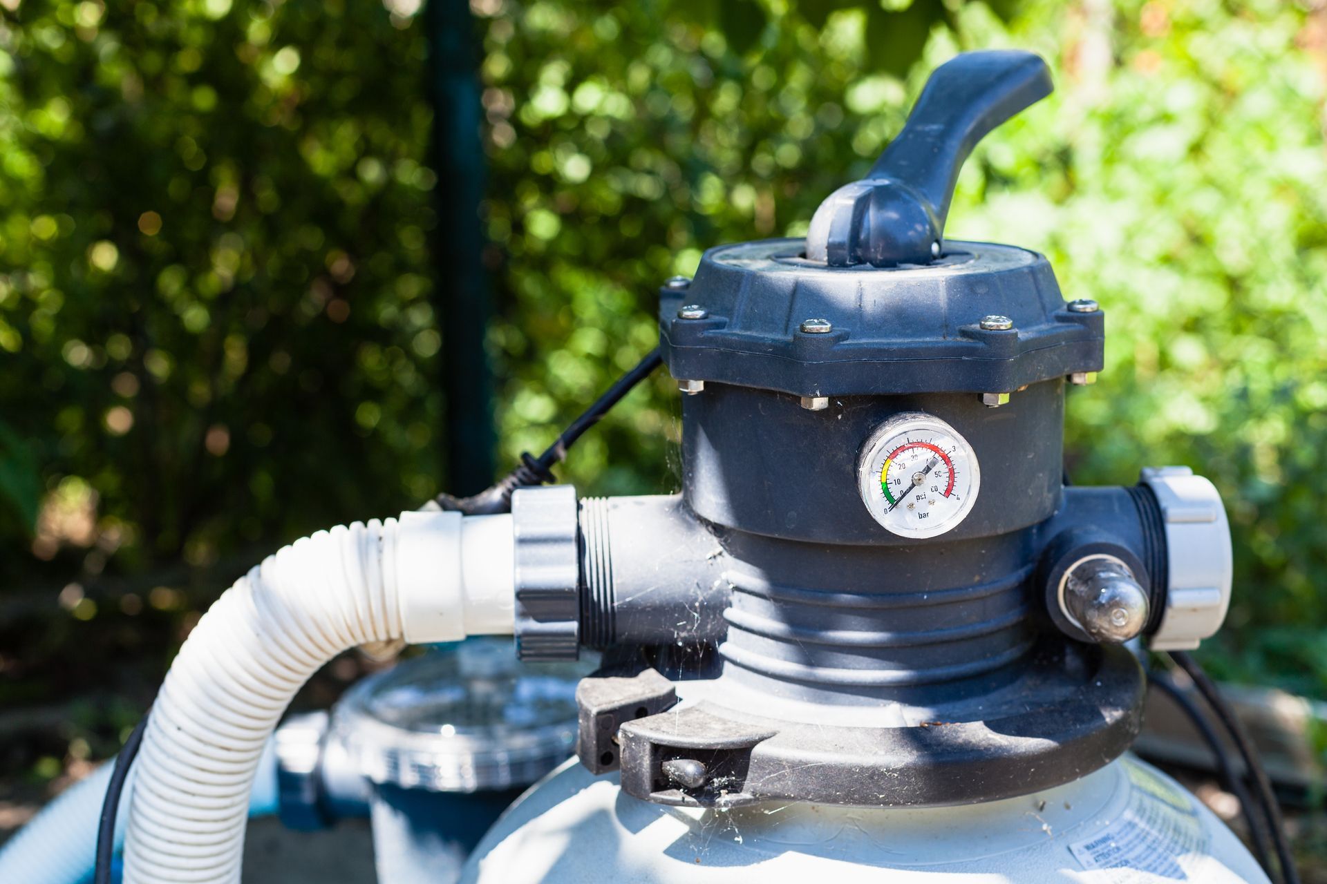 A dark grey pool filter head with a pressure gauge and valve handle, positioned outdoors against a blurred green background.