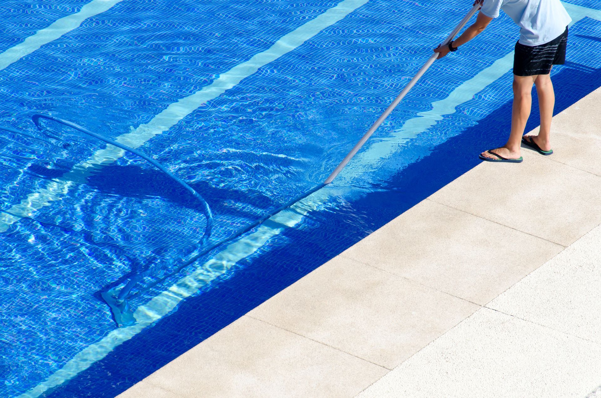 A person uses a long-handled net to clean debris from a bright blue swimming pool on a sunny day.