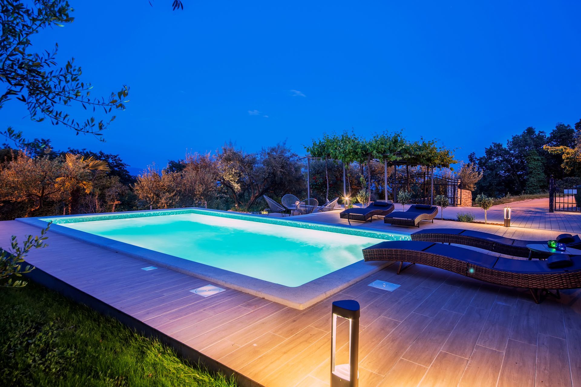 A bright, illuminated swimming pool on a wooden deck at dusk, surrounded by trees under a deep blue sky.
