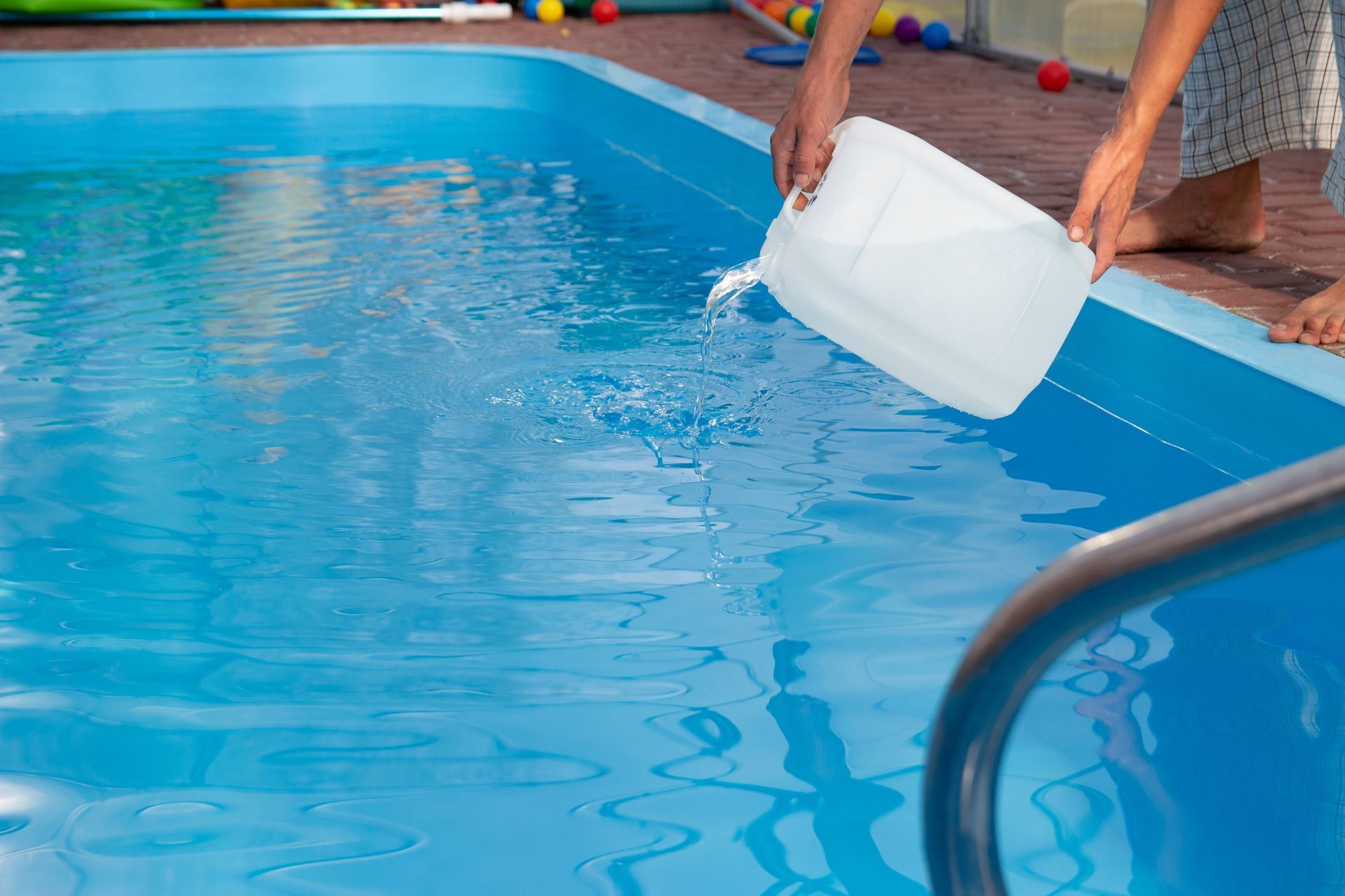 A person pours a clear liquid from a white plastic container into a bright blue swimming pool.