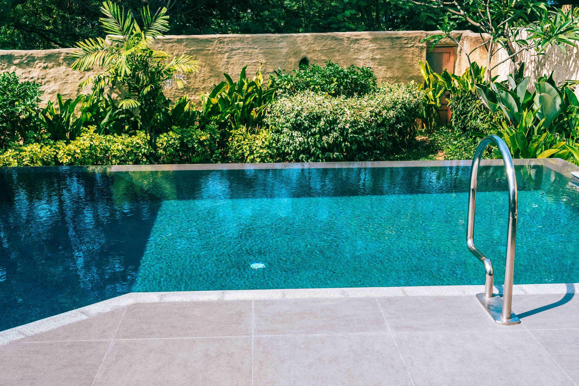 Outdoor swimming pool with clear blue water, greenery behind it, and a metal handrail at the edge