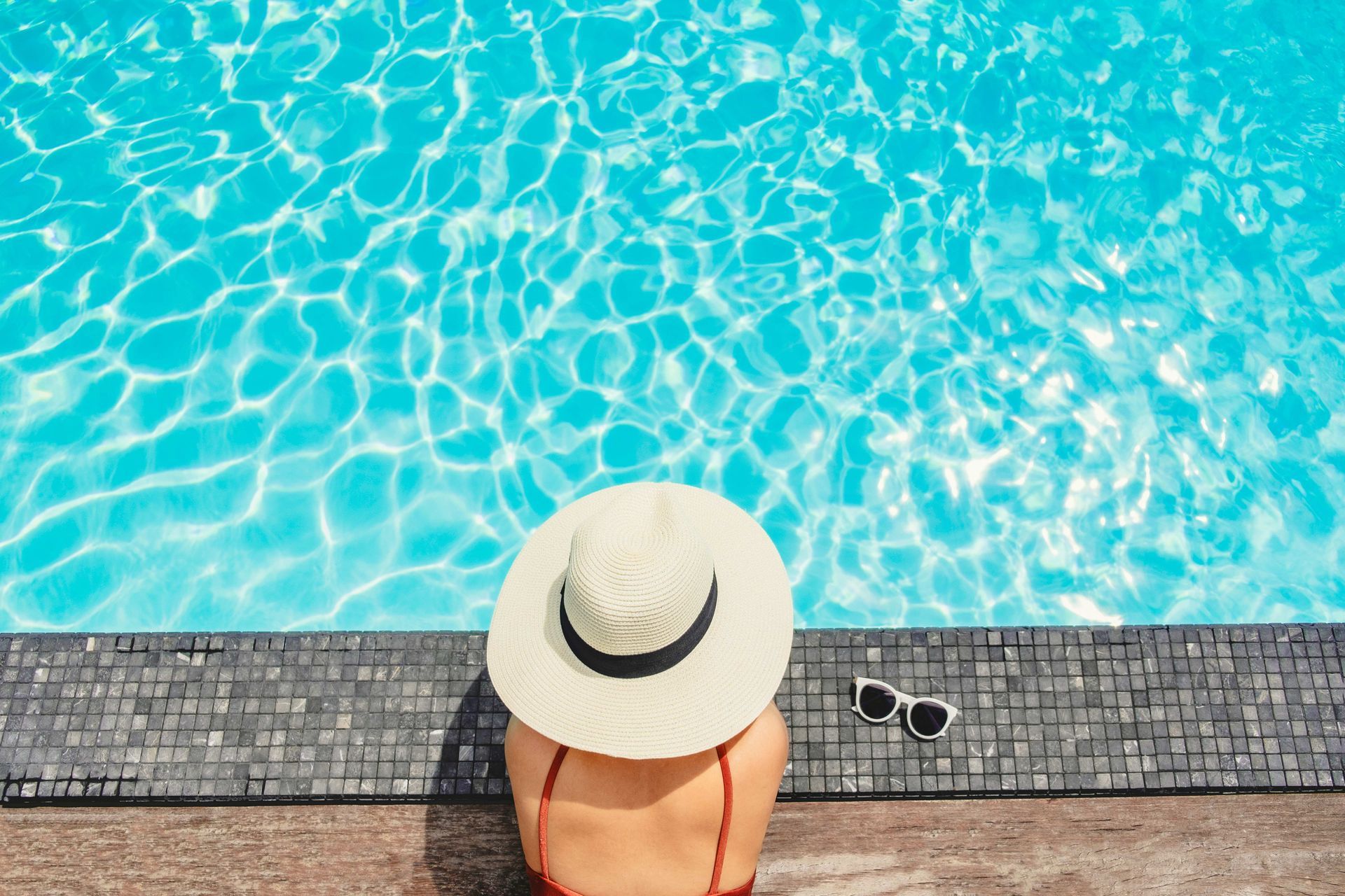 Person in a white sunhat sitting by a bright turquoise swimming pool, with sunglasses on the deck.
