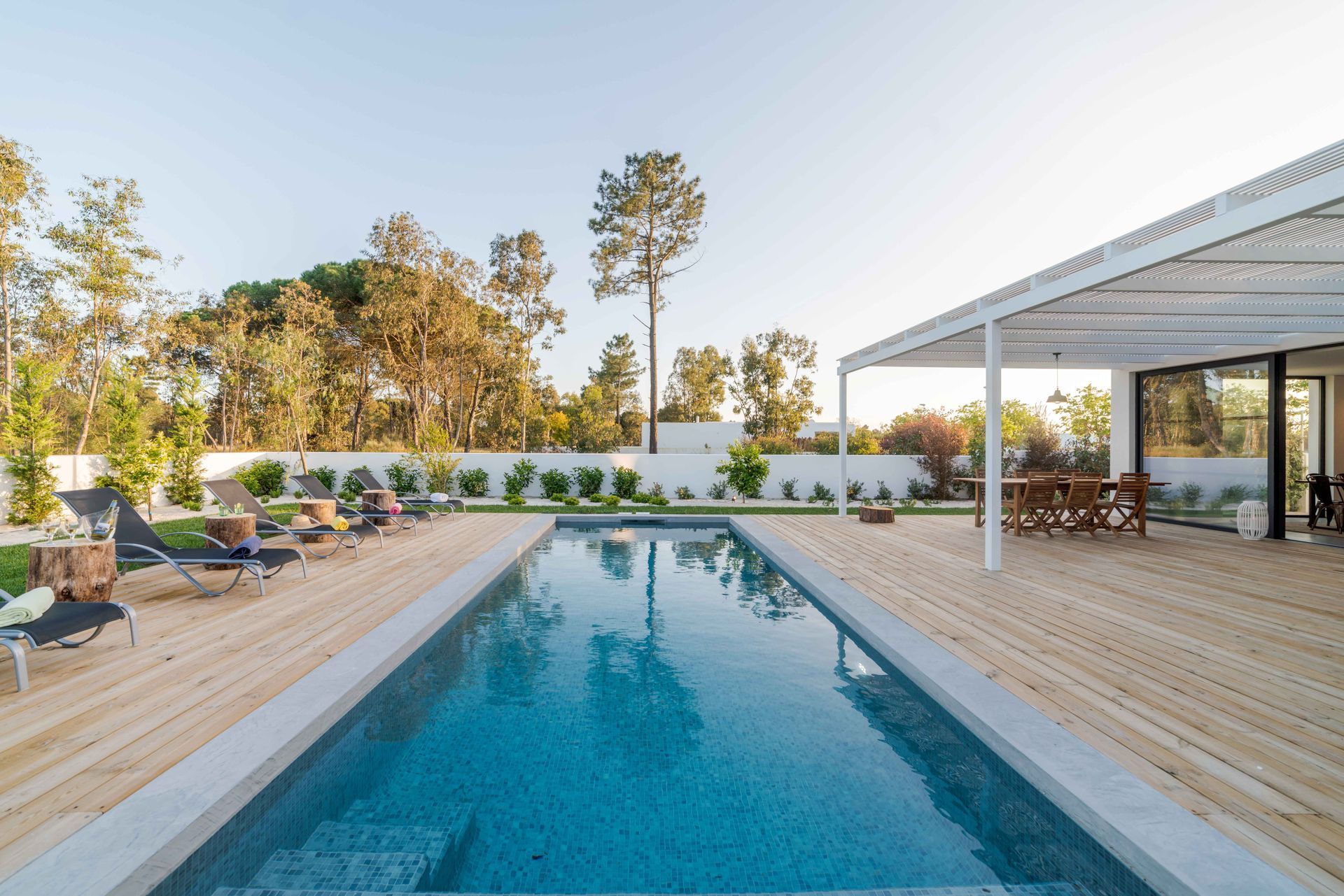 Backyard pool with wooden deck, lounge chairs, and a covered patio at sunset