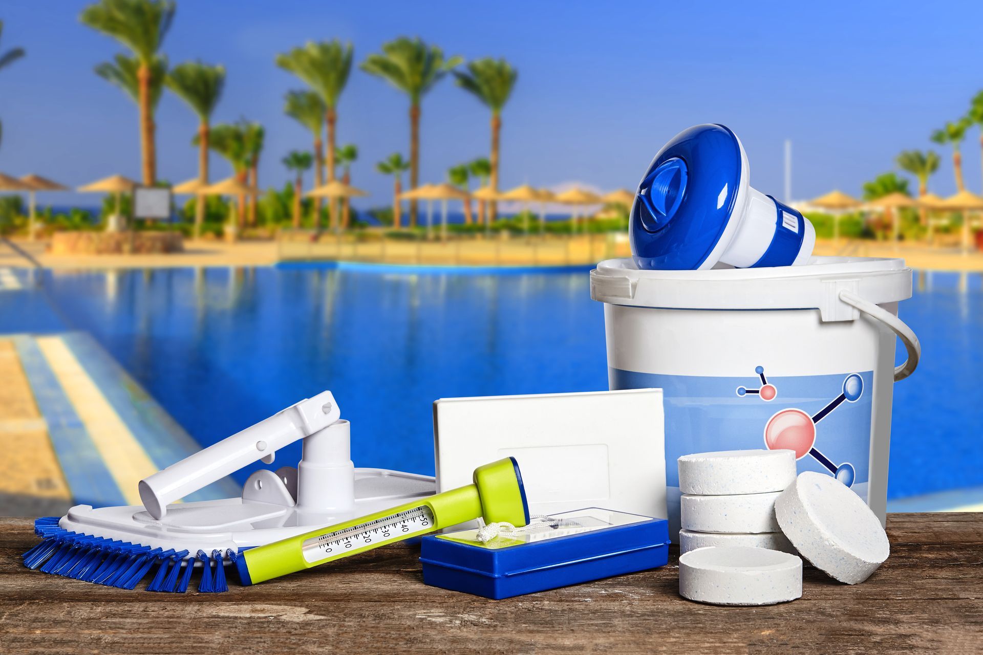 Pool cleaning supplies, including a brush, thermometer, and chlorine tablets, sit on a surface with a pool in the background.