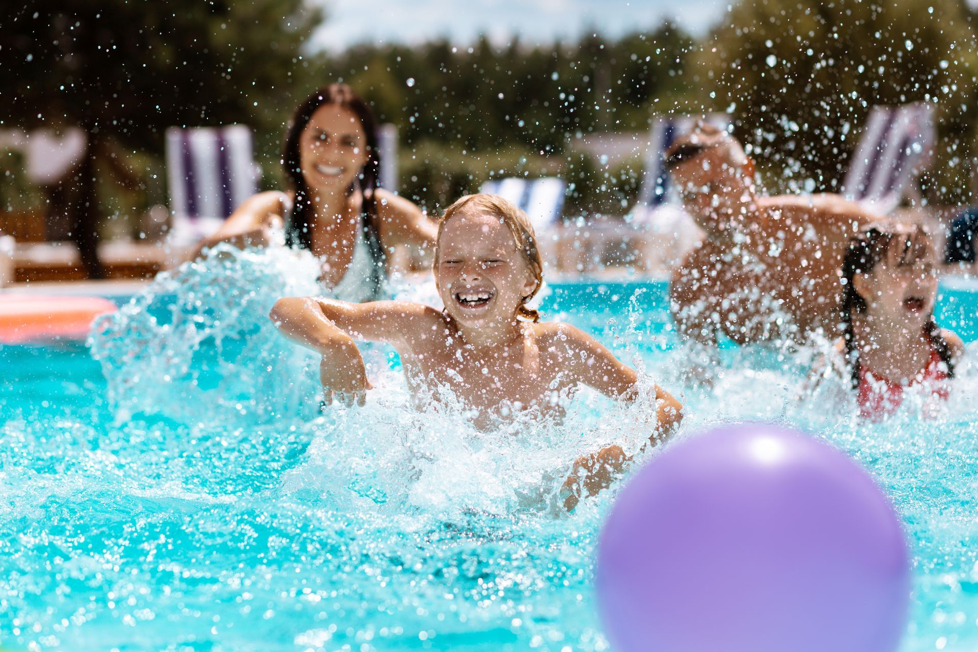 A happy family splashing in a sunny swimming pool with a purple ball in the foreground.