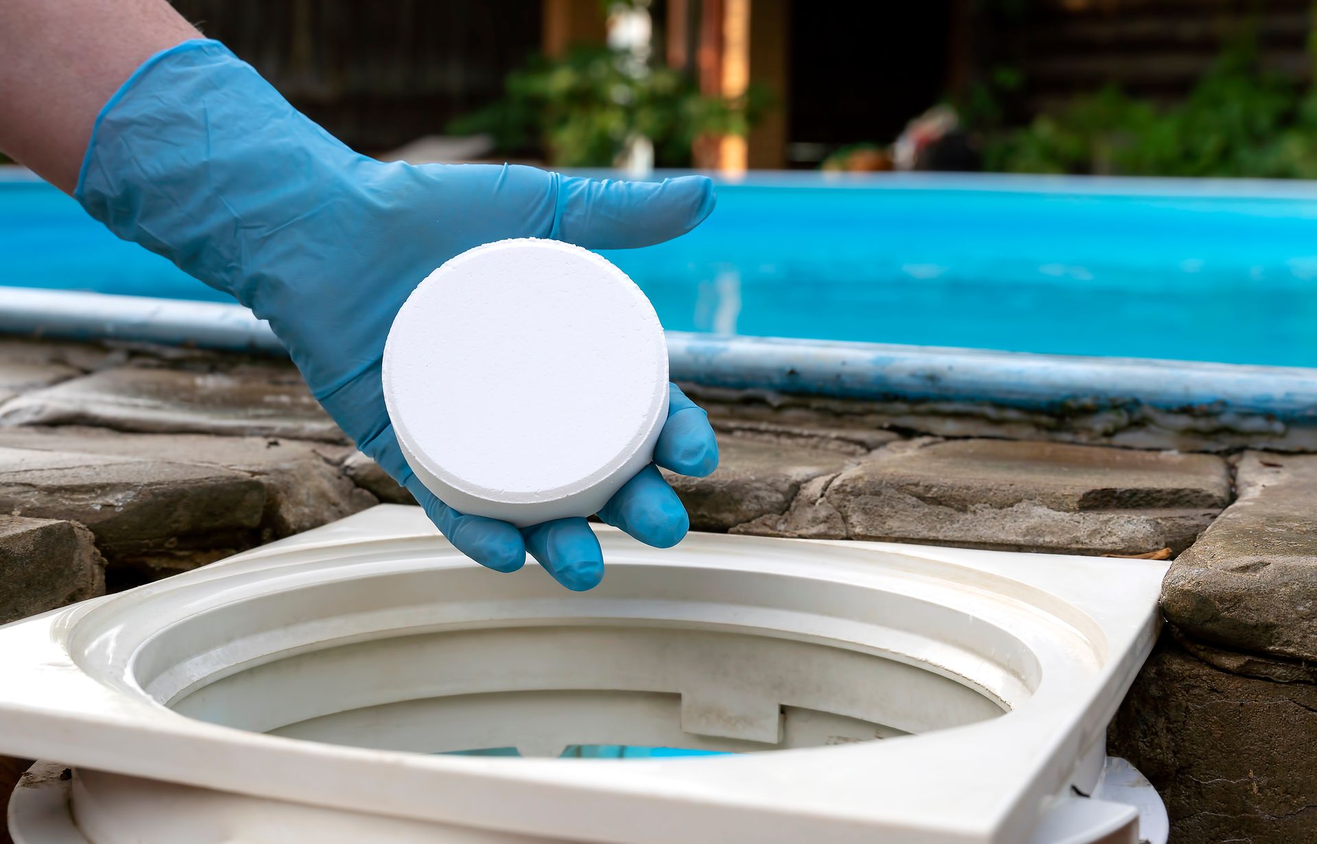 A gloved hand holds a round white pool chemical tablet over a pool skimmer basket.