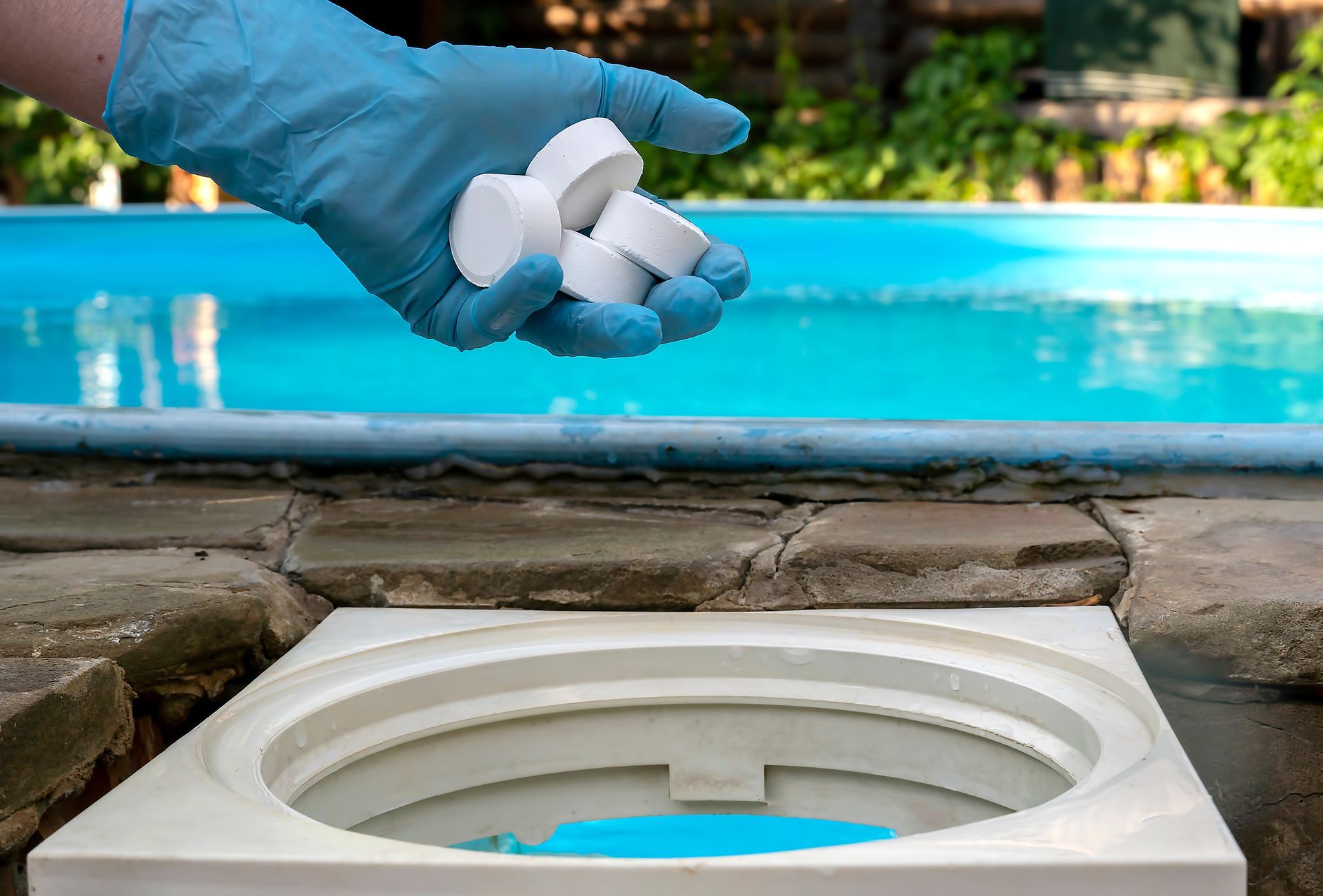 A gloved hand holds pool chlorine tablets over a skimmer basket near a blue swimming pool.