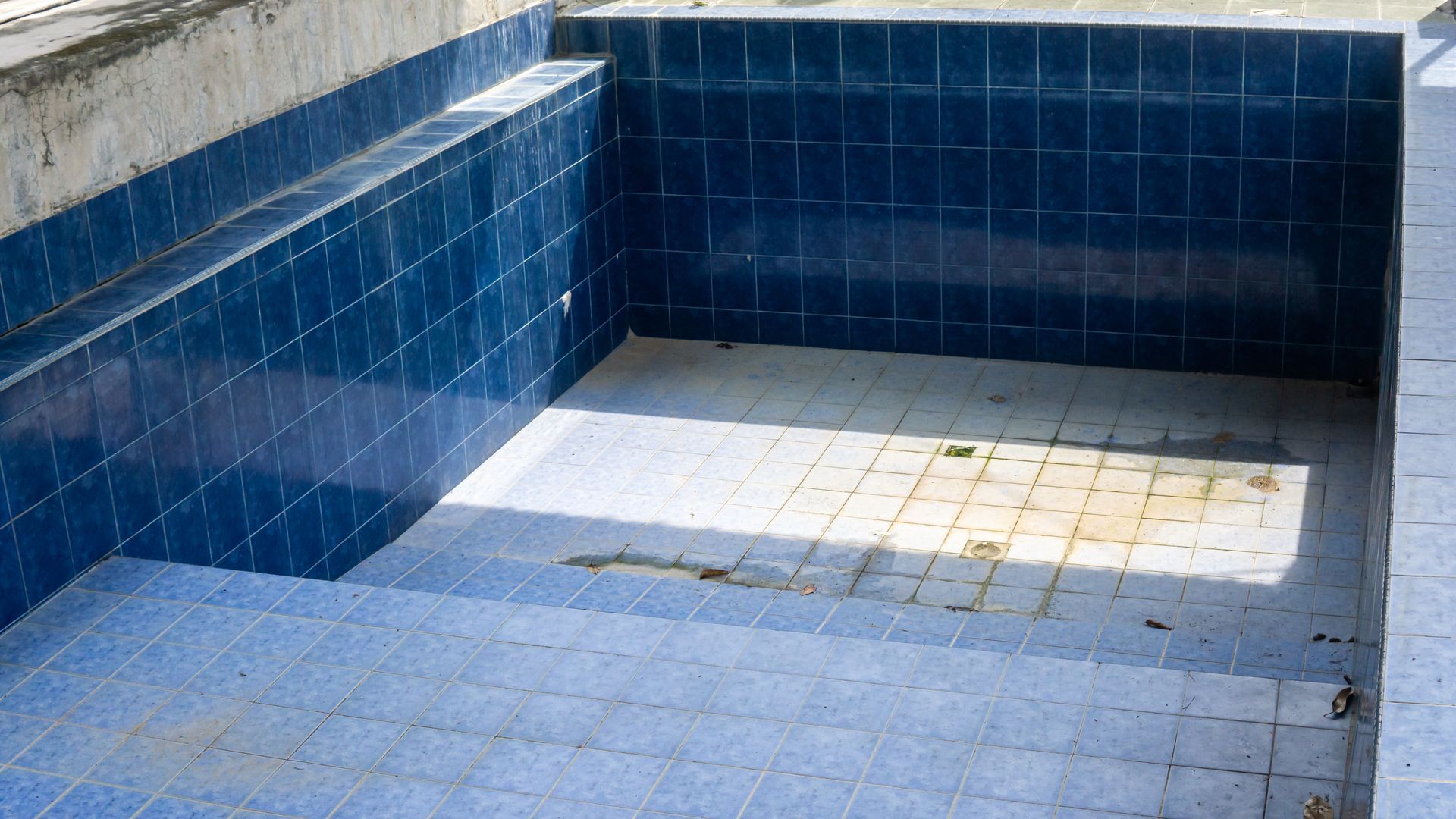 A close-up view of an empty rectangular swimming pool lined with dark blue tiles and a light-colored tiled floor.