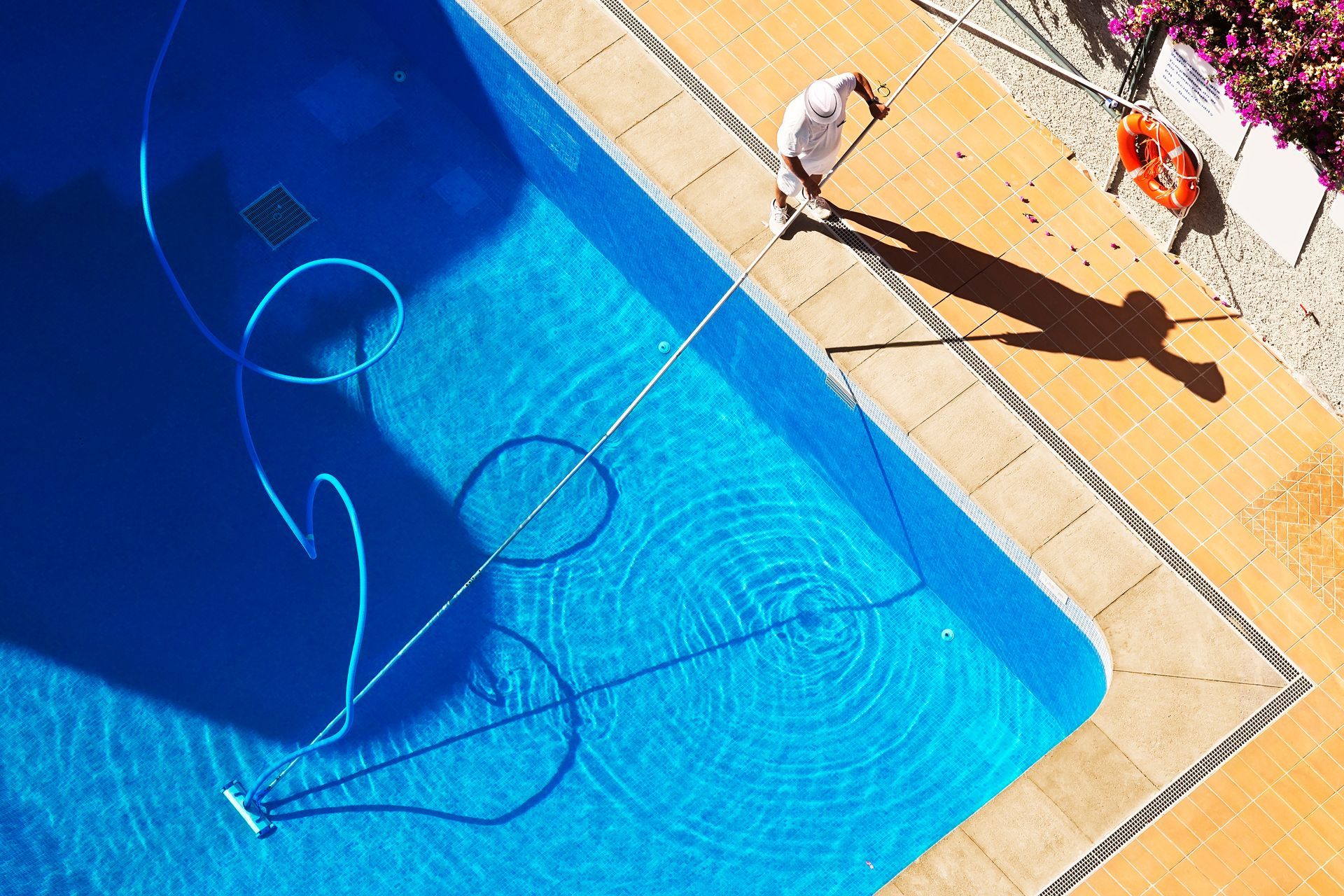 A top-down view of a person using a long-handled pole to clean a bright blue swimming pool on a sunny day.