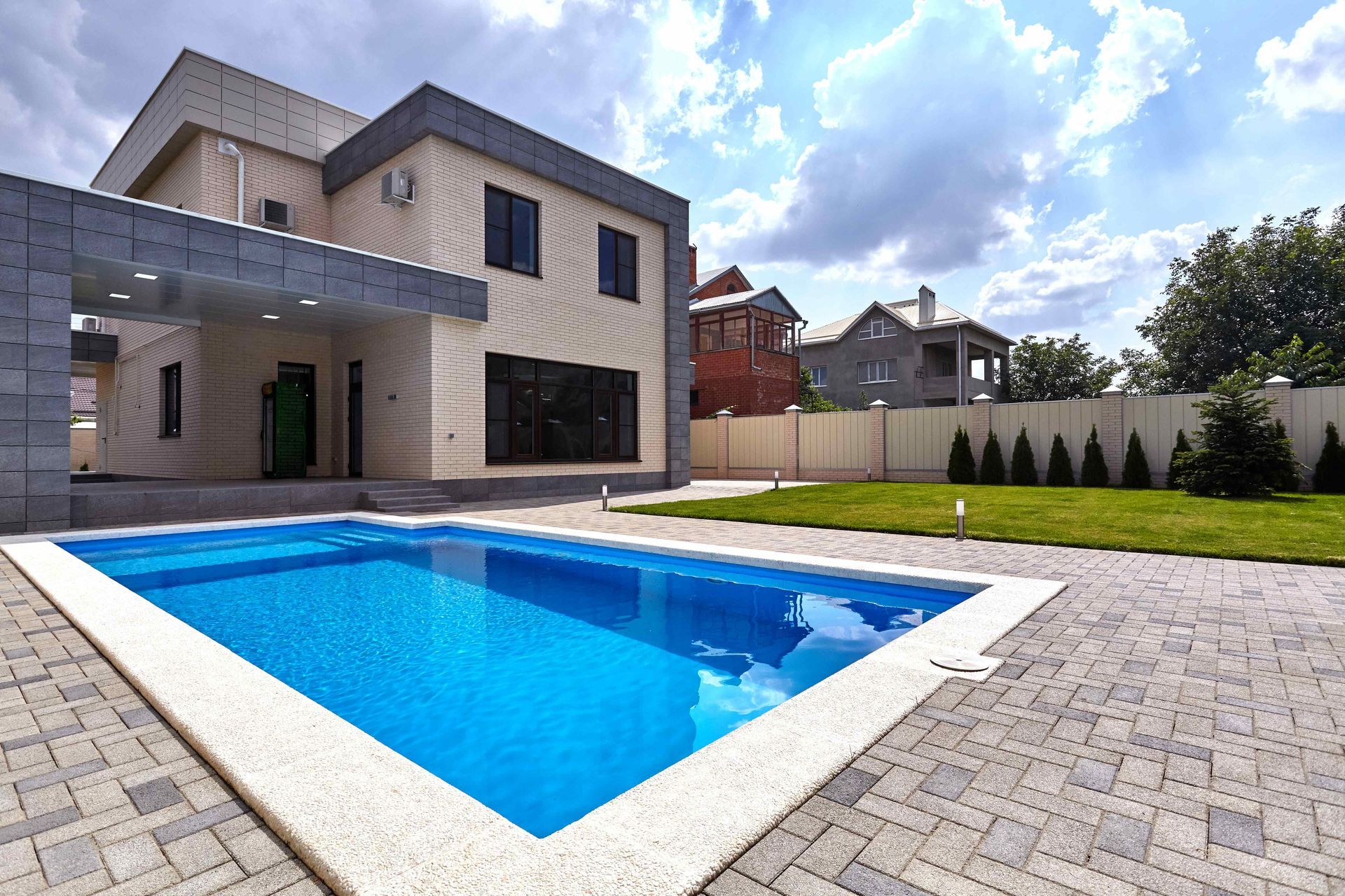 Modern two-story house with a blue swimming pool and paved backyard patio under a partly cloudy sky