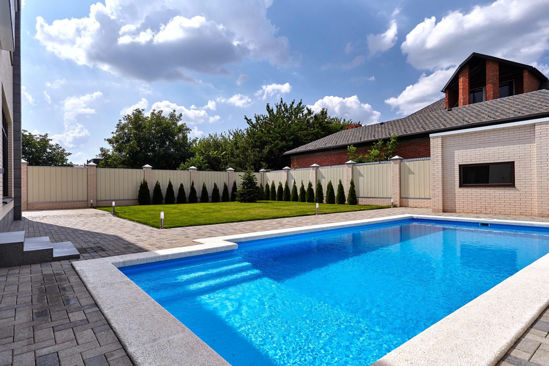 Backyard with a blue swimming pool, stone patio, and modern house under a partly cloudy sky