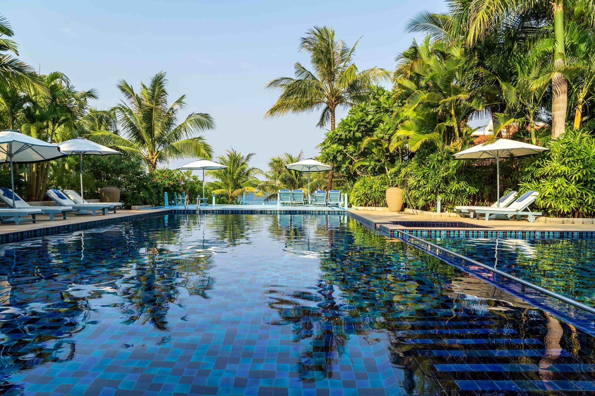 Tropical resort pool with blue tile water, palm trees, and lounge chairs under a bright sky