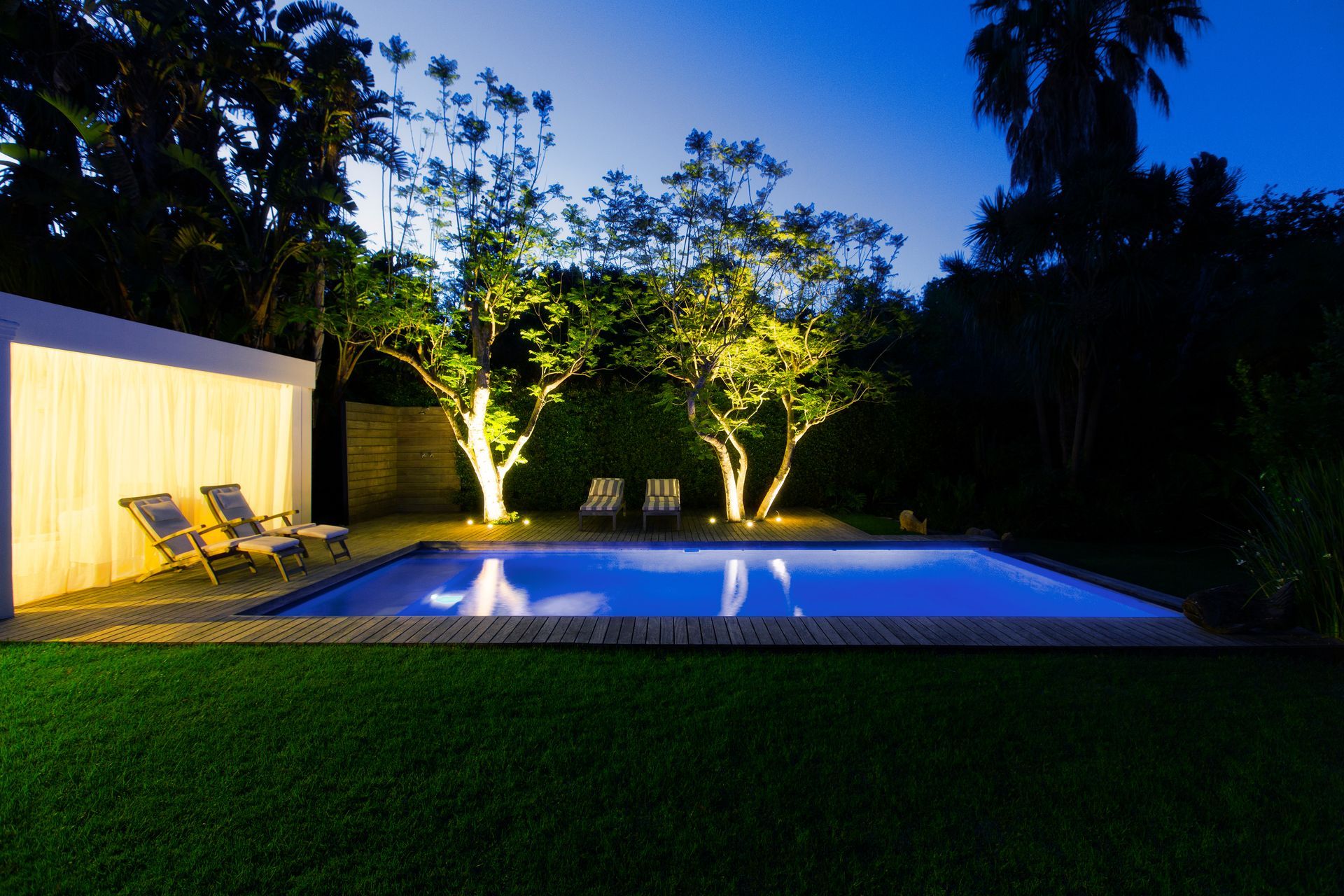A swimming pool at night, illuminated with blue water, glowing trees, and lounge chairs on a grassy lawn.