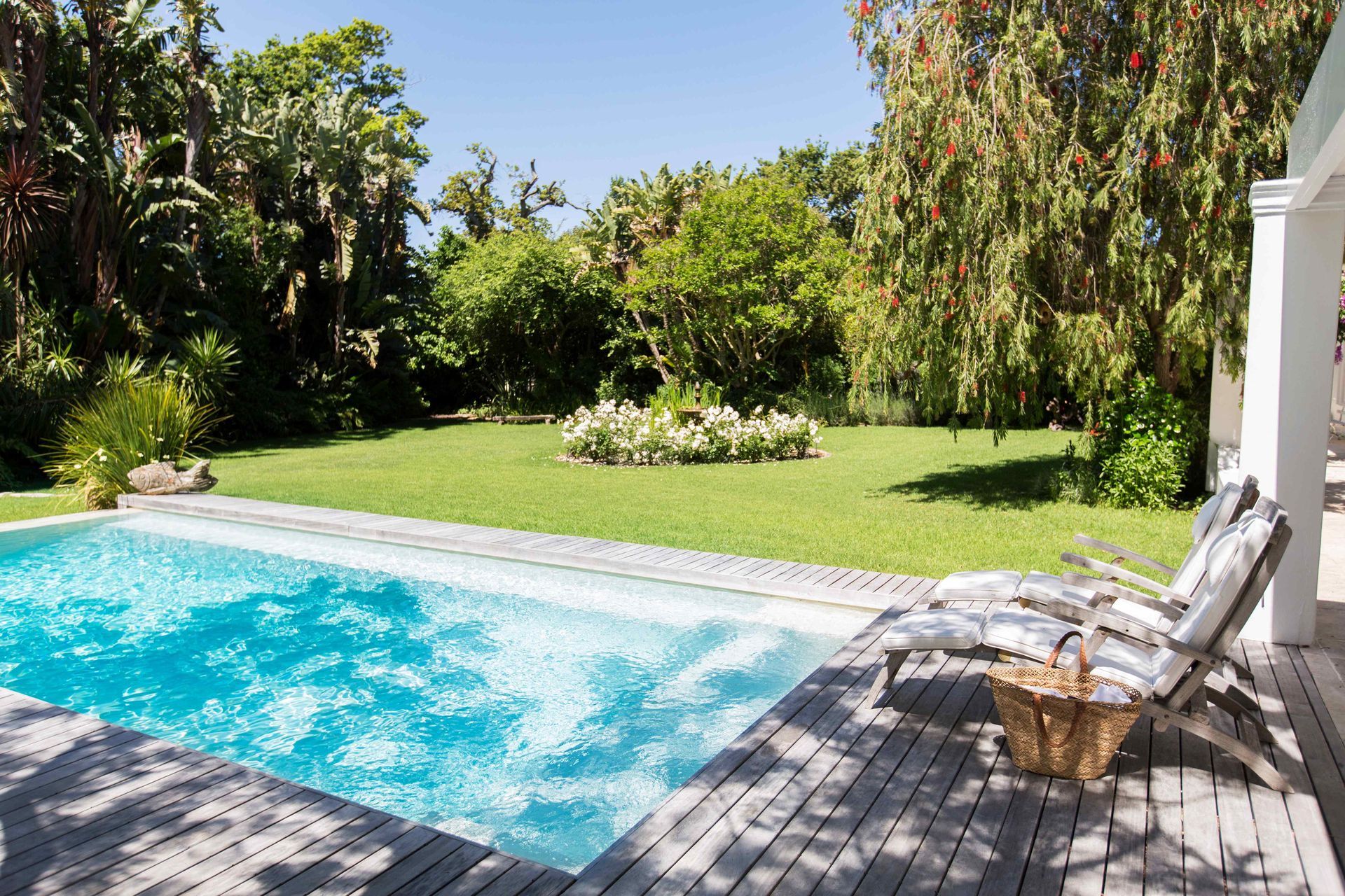 Backyard pool beside a wooden deck, with lounge chairs and lush green trees in bright sunlight