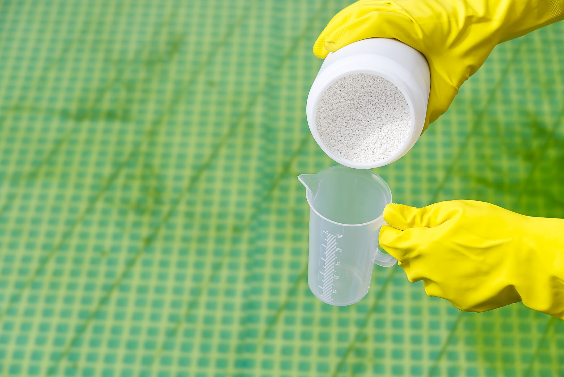 Hands in yellow gloves pouring white granules from a container into a measuring cup against a green grid background.