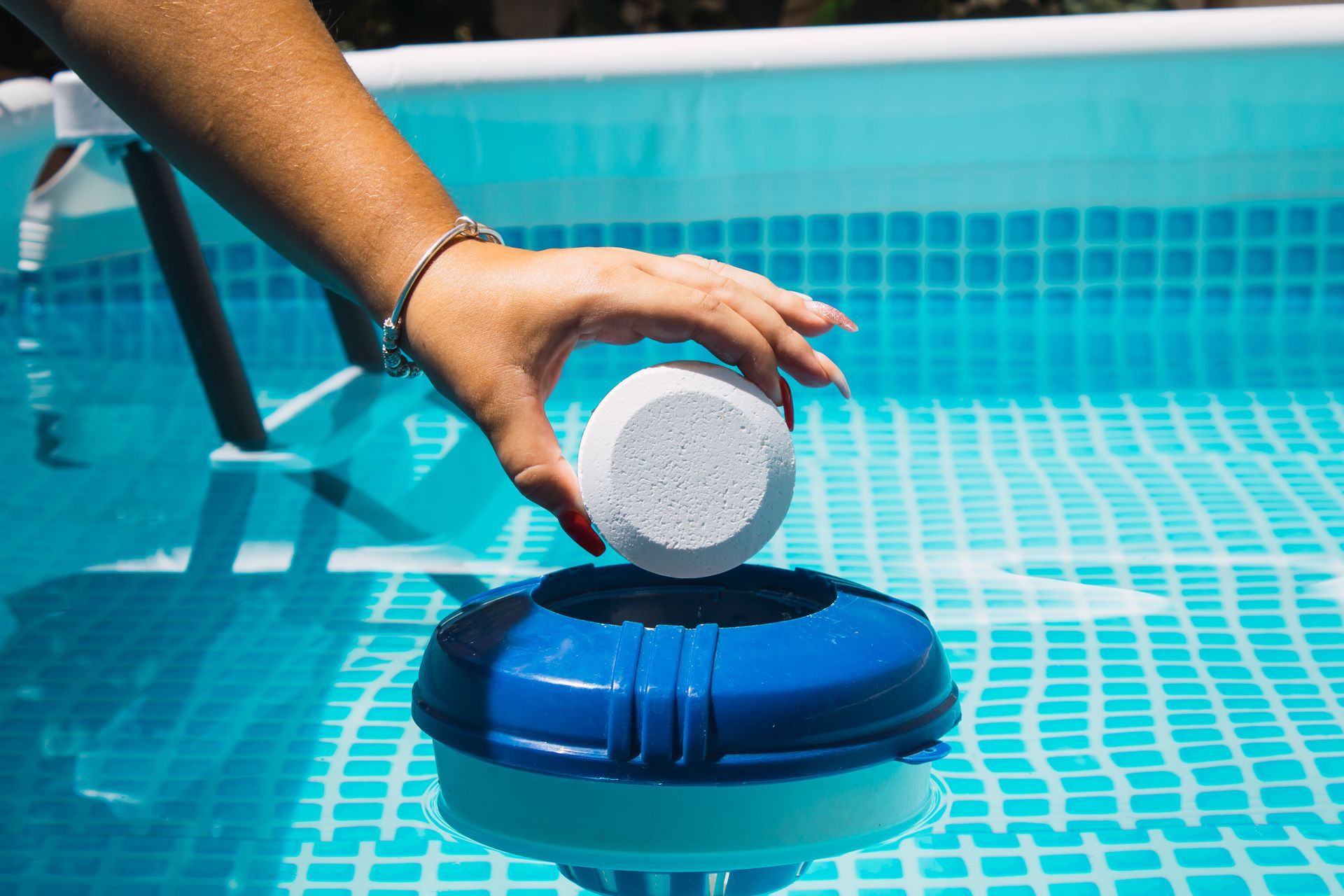 A hand places a white chlorine tablet into a blue floating pool dispenser in a bright blue swimming pool.