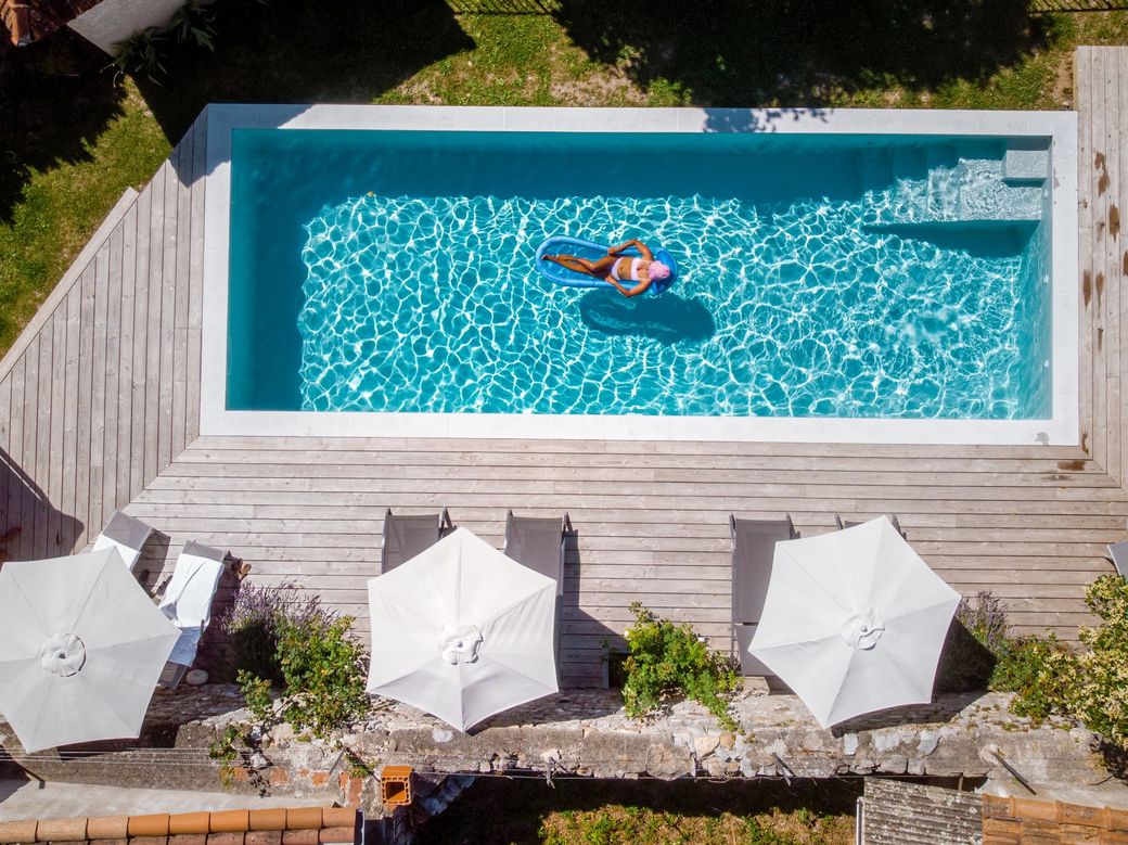 An aerial view of a rectangular swimming pool with a person on a blue float, surrounded by white umbrellas and a deck.
