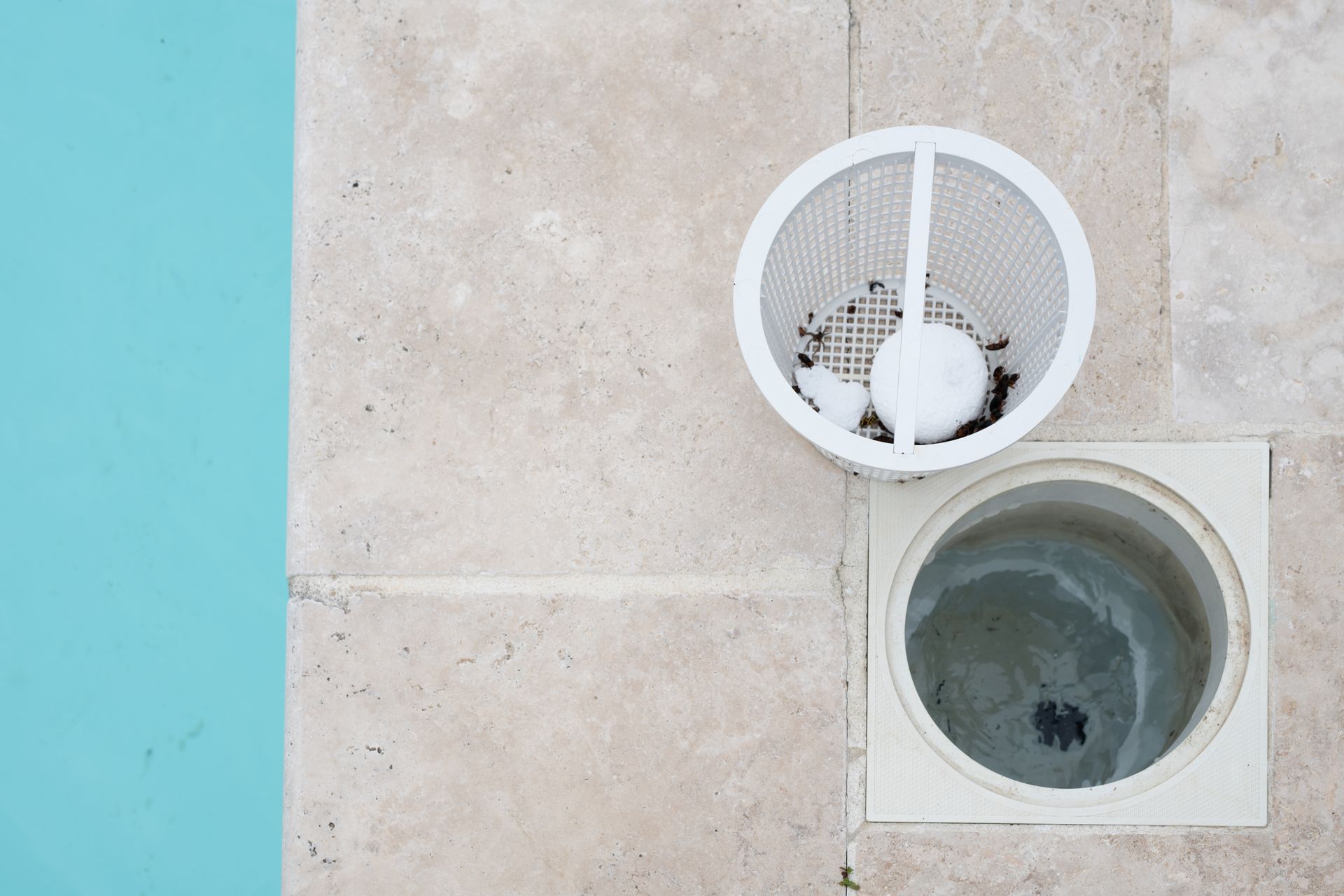 A white pool skimmer basket sitting on a stone deck next to an open pool skimmer well and blue water.