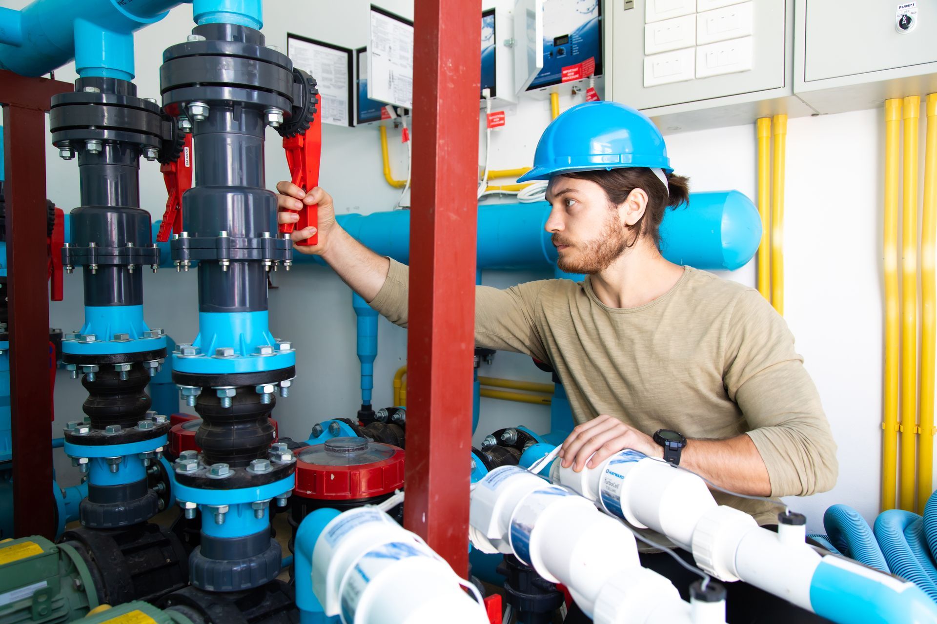 A technician in a blue hard hat operates a red valve on a large blue industrial pipe system in a utility room.