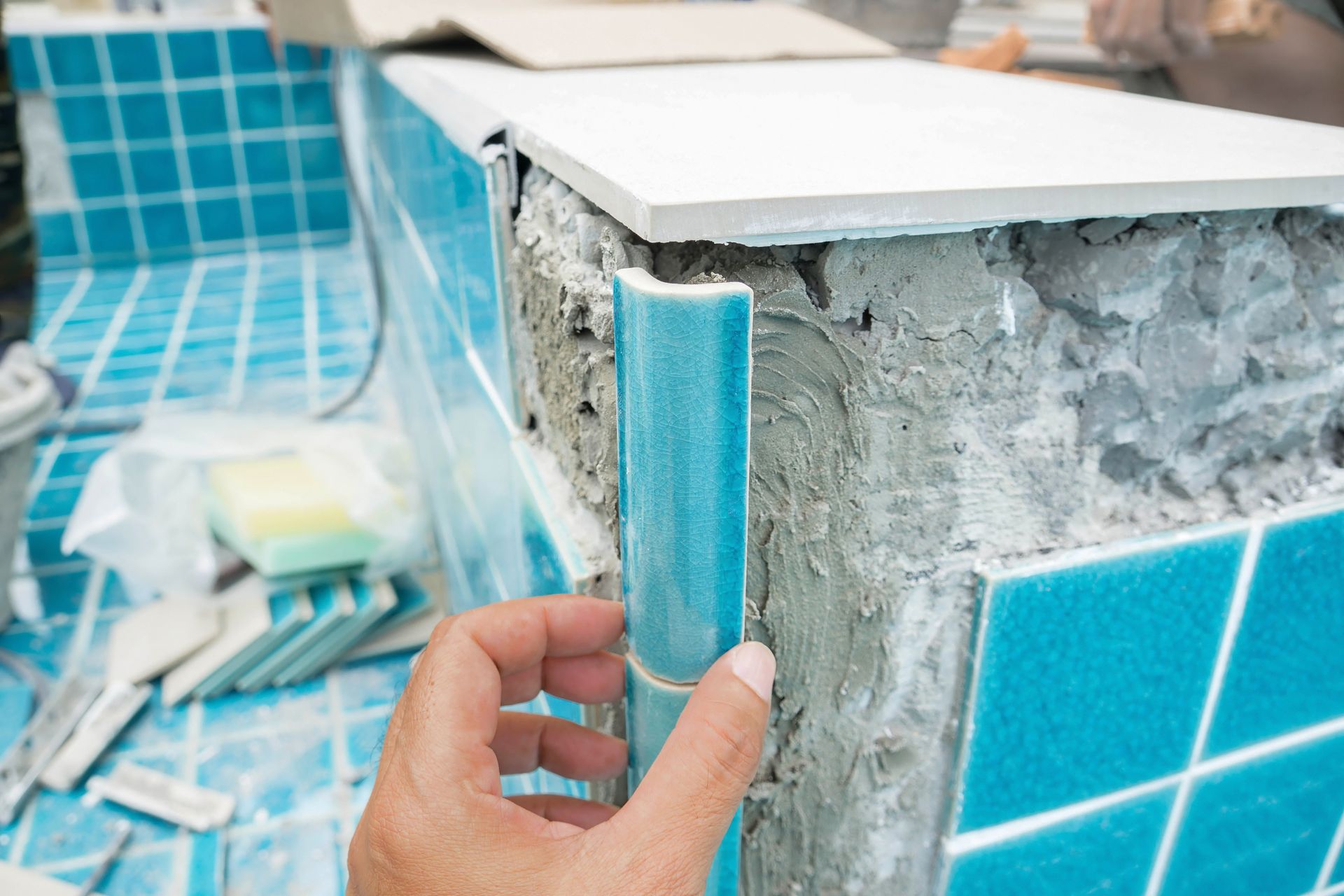 A hand holds a curved teal edge tile against the concrete corner of a swimming pool undergoing repair.