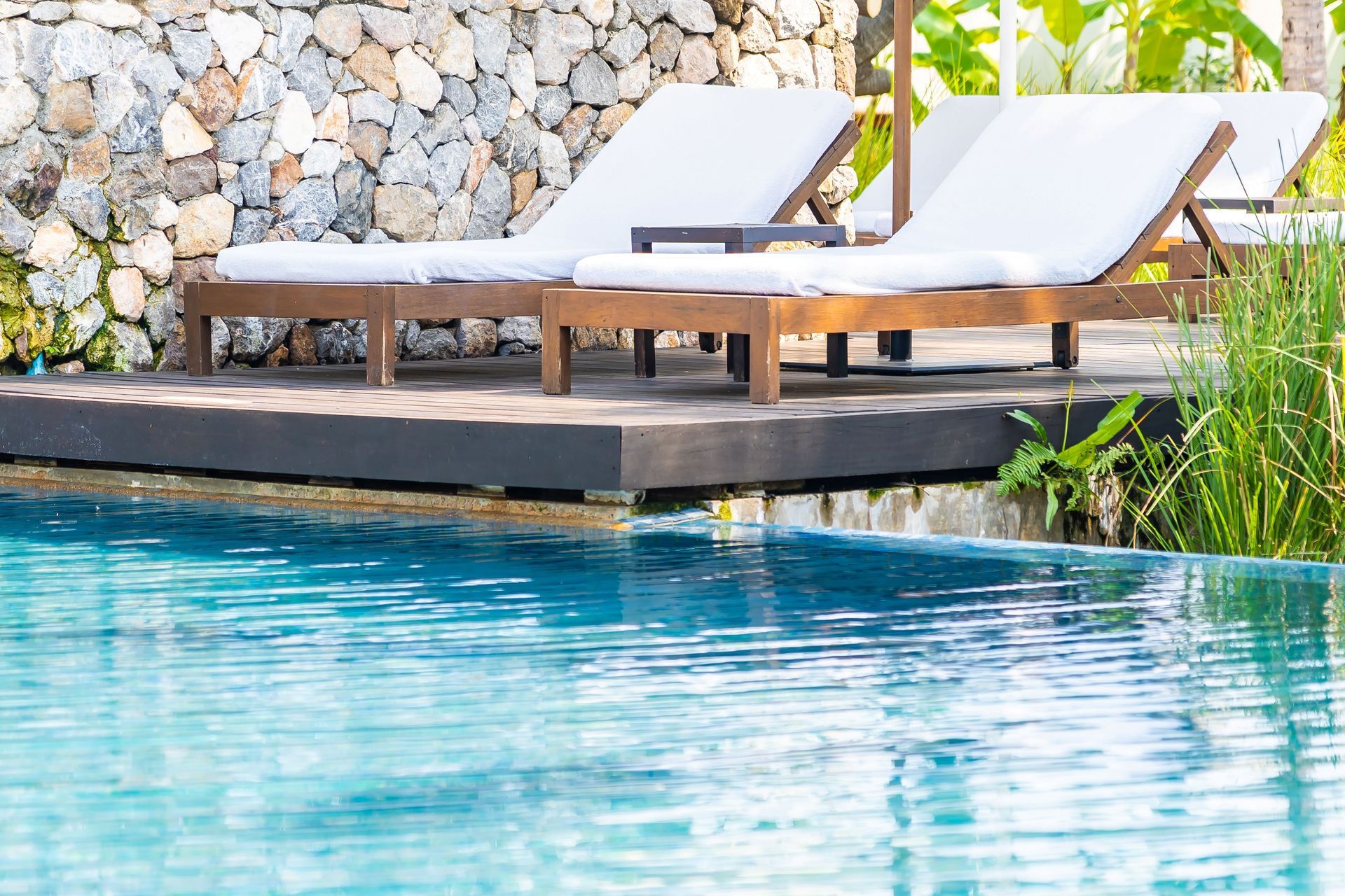 Poolside lounge chairs beside a turquoise swimming pool and stone wall