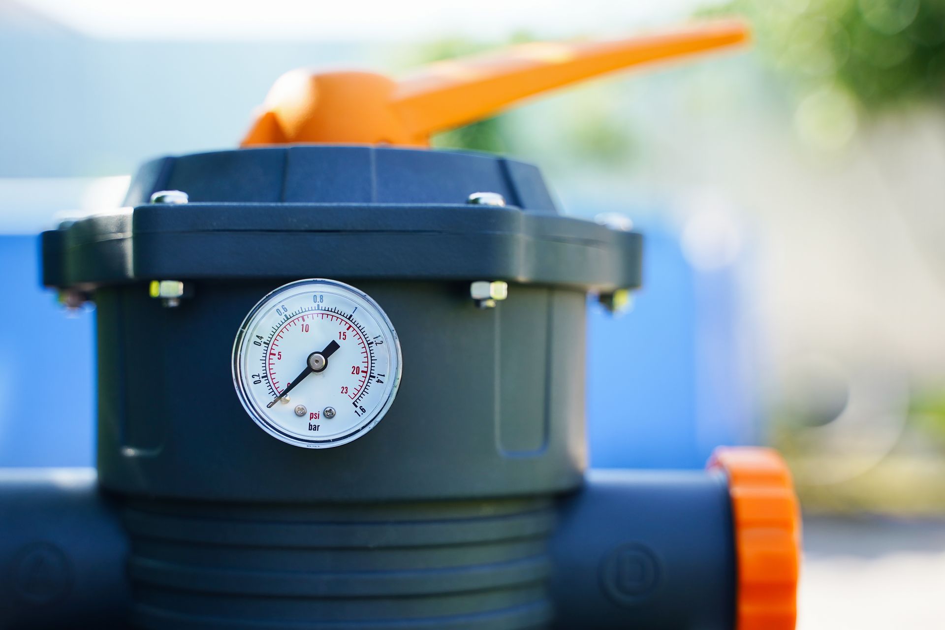 A dark gray pool filter valve with an orange handle and a pressure gauge set to low, displayed against a blurred background.