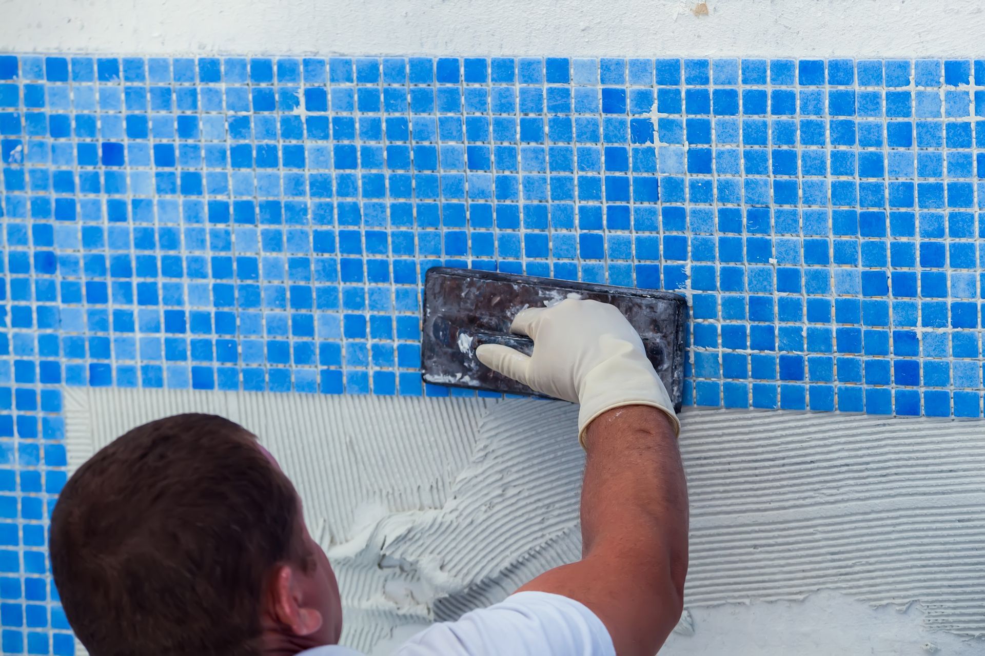 A worker wearing a protective glove spreads adhesive onto a wall to install small blue mosaic tiles.