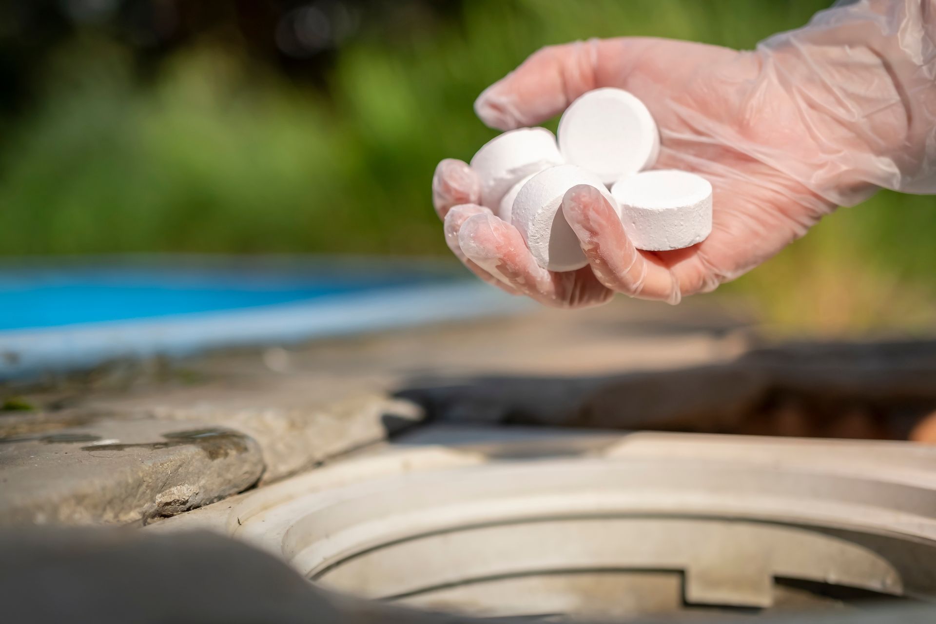 A gloved hand holding several white chlorine tablets above an open swimming pool skimmer.