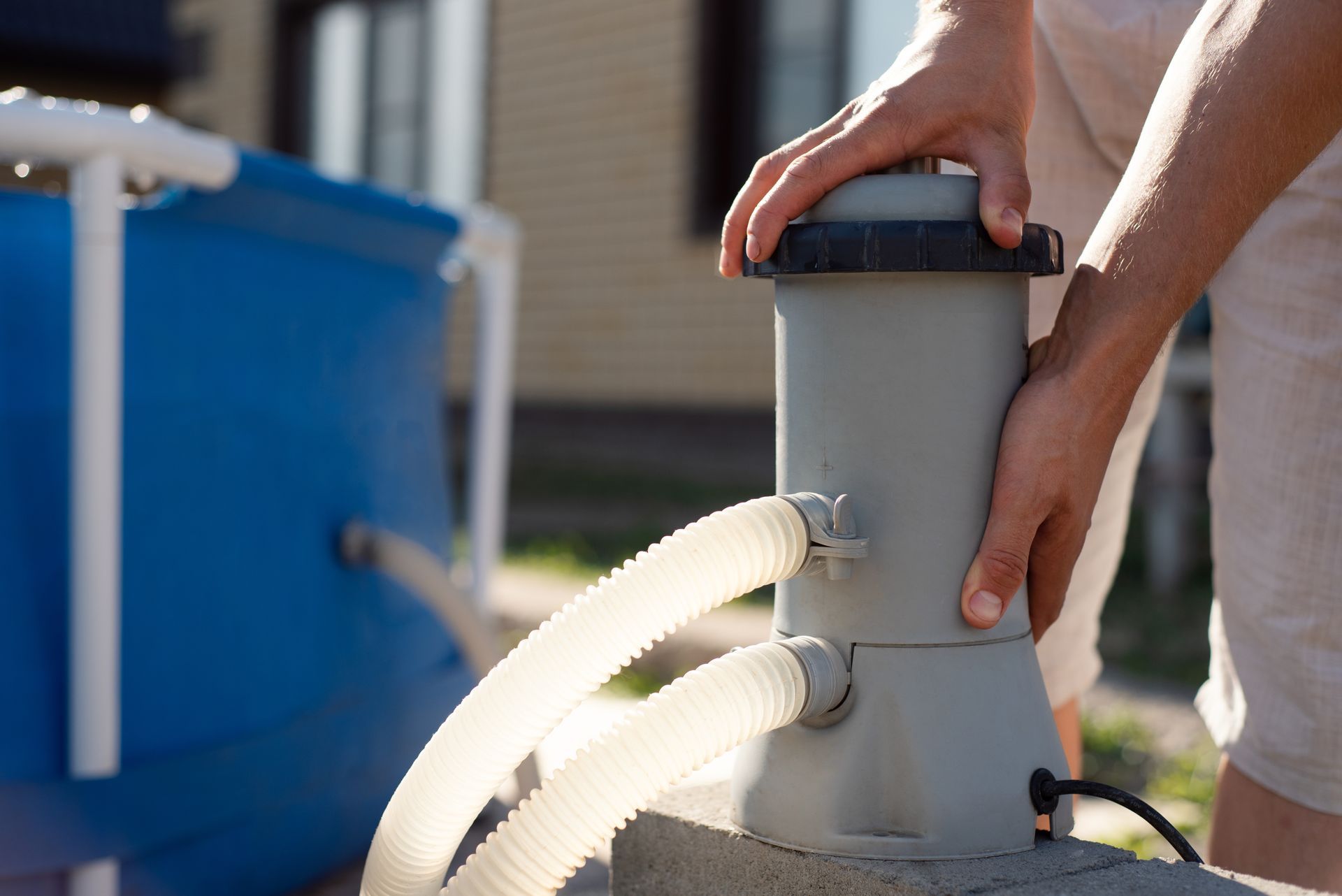A person sets up a grey pool filter pump connected by hoses to a blue frame pool outdoors.