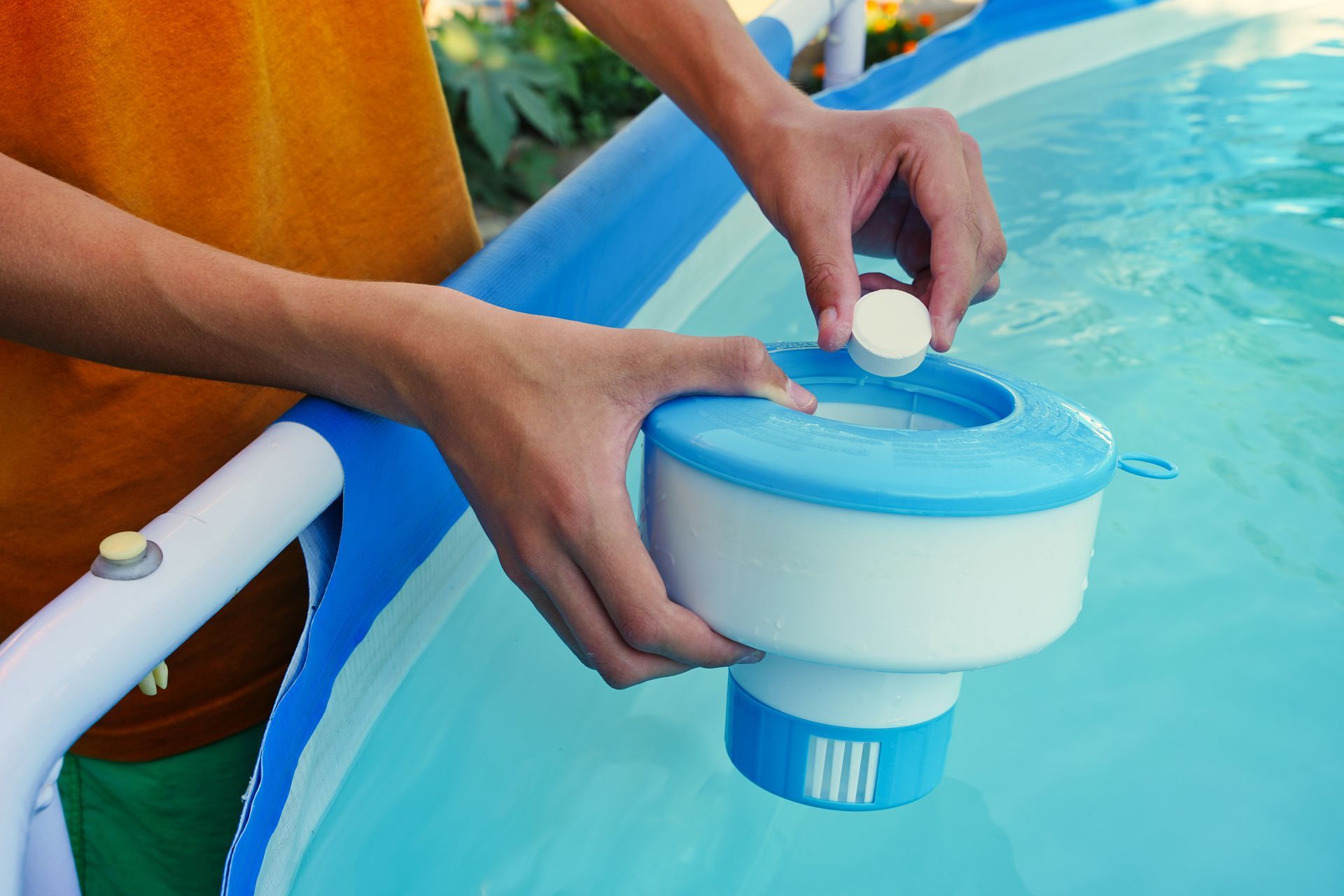 A person placing a small chlorine tablet into a floating pool chemical dispenser above a blue above-ground swimming pool.
