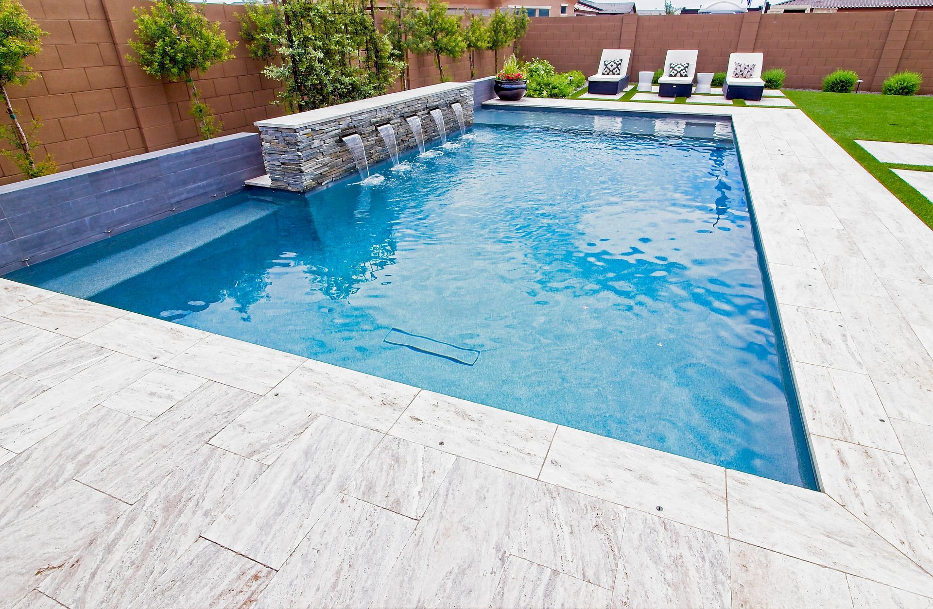 A rectangular backyard swimming pool with a stone water feature, light tile decking, and lounge chairs on a grassy lawn.