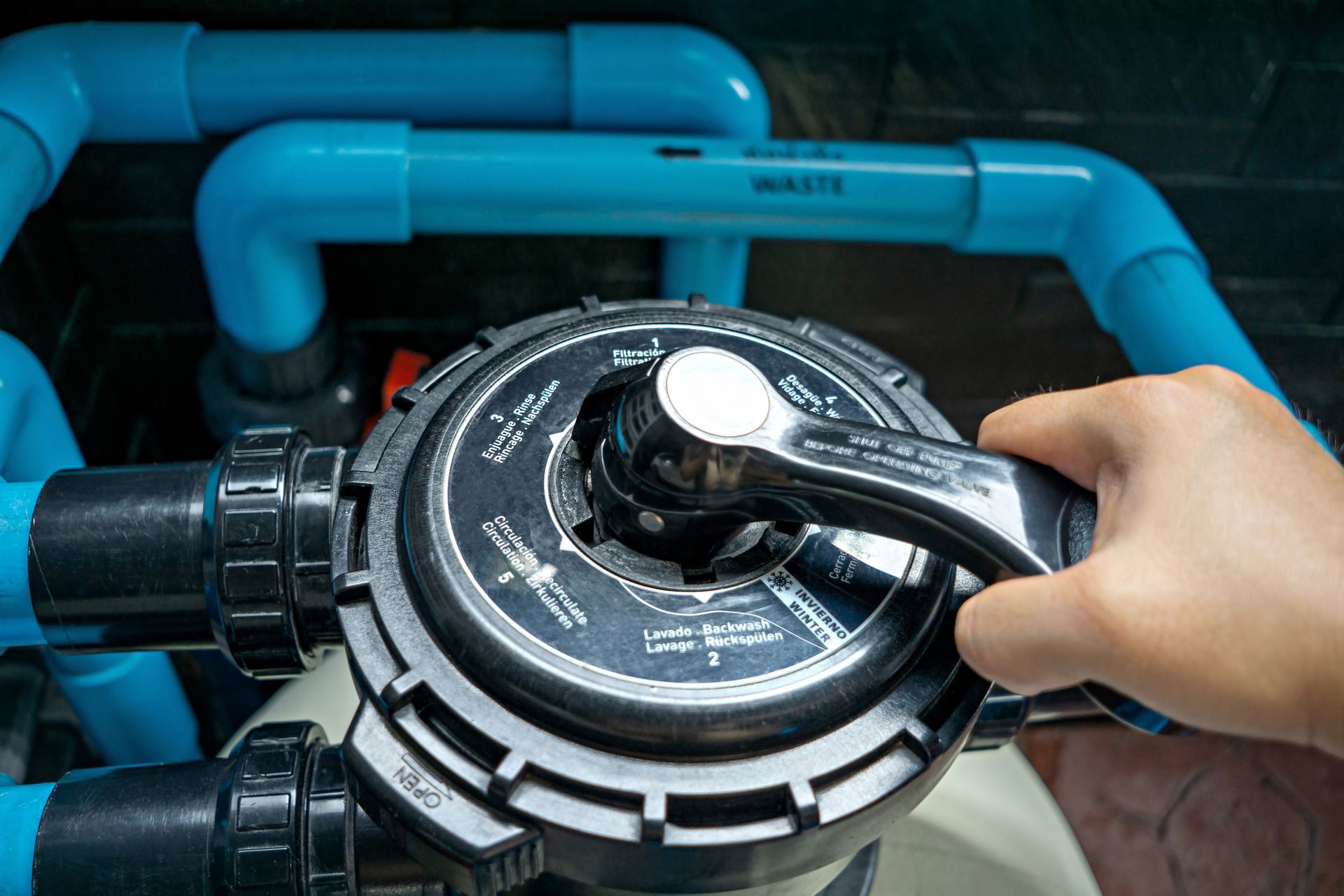 A hand turns the multiport valve handle on a pool sand filter, with blue PVC pipes visible in the background.