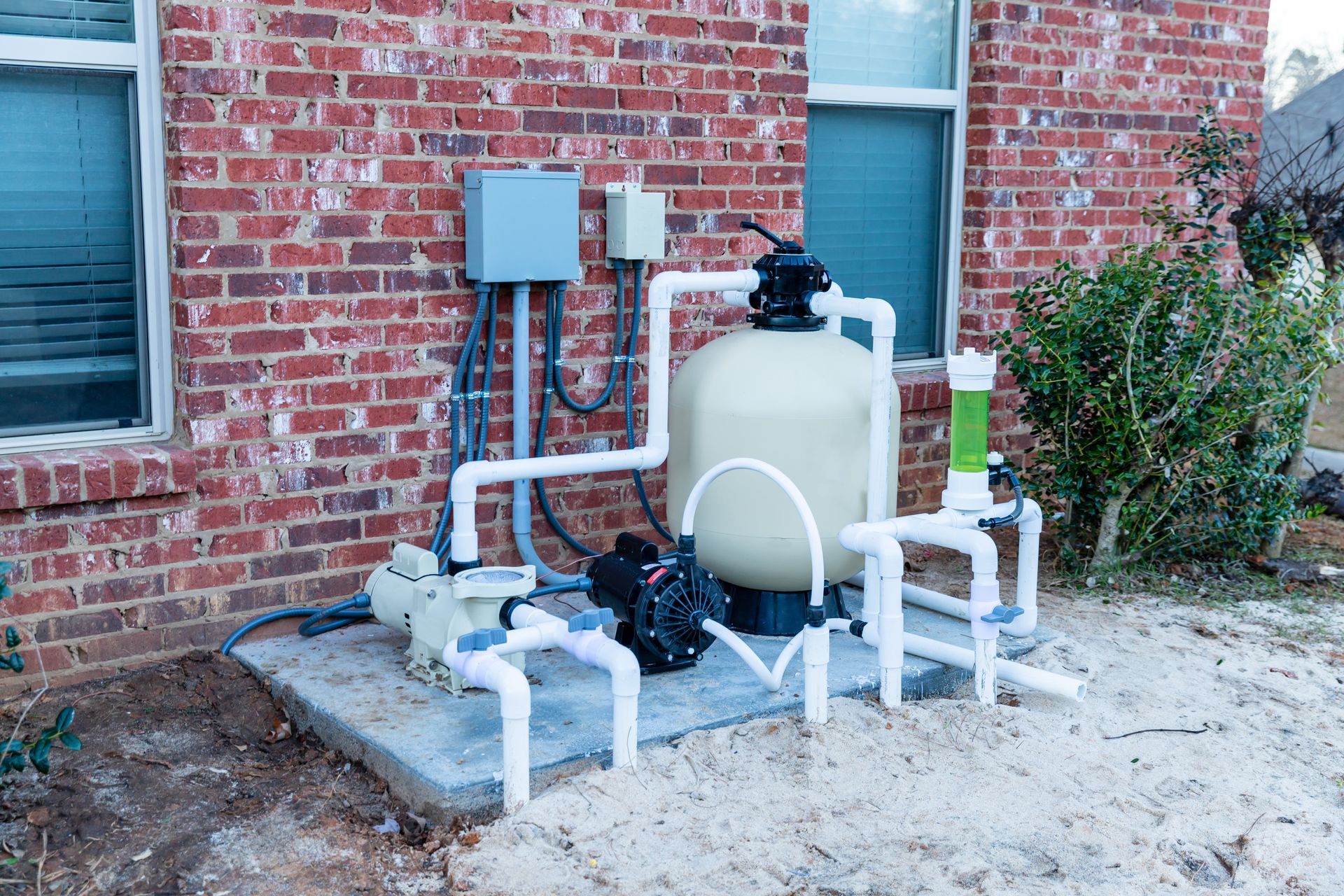 A swimming pool pump and filtration system sits on a concrete pad outside a brick house near a green shrub.