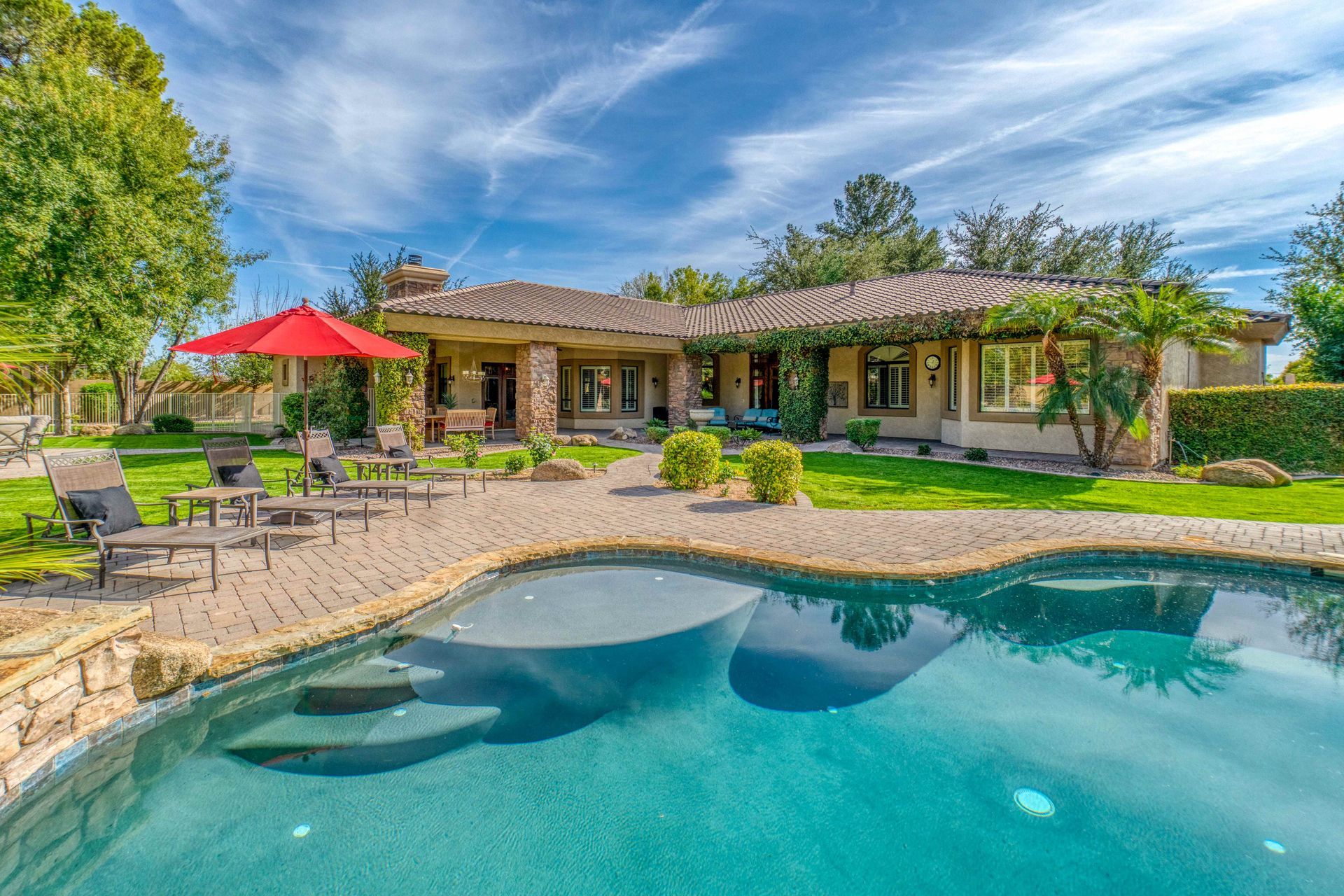Backyard pool with stone patio, lounge chairs, red umbrella, and a single-story house in the background