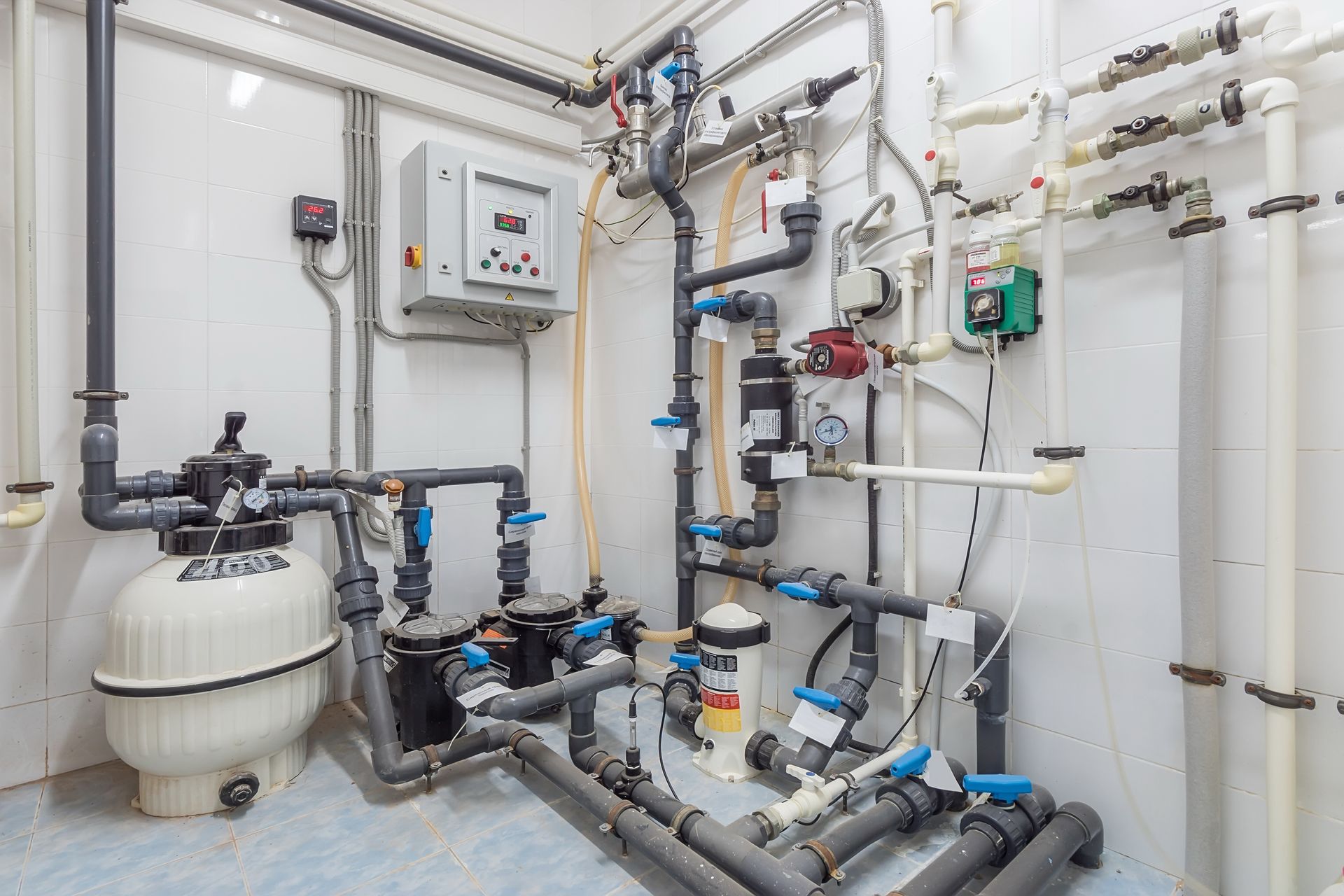 A swimming pool filtration and pump system with various pipes, valves, and a control box mounted on a white wall.