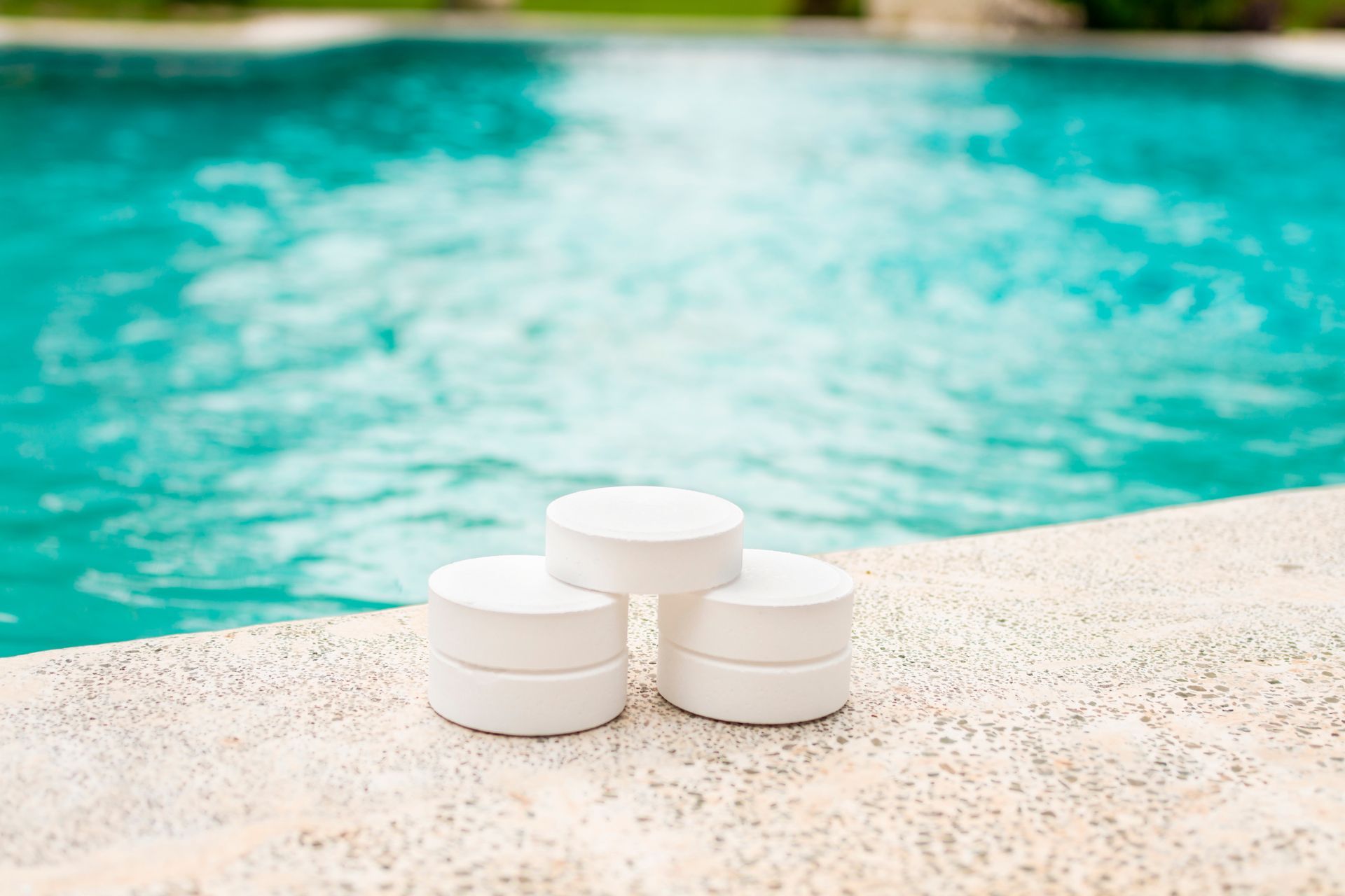 Three white pool chemical tablets stacked on a textured pool deck edge overlooking a turquoise swimming pool.