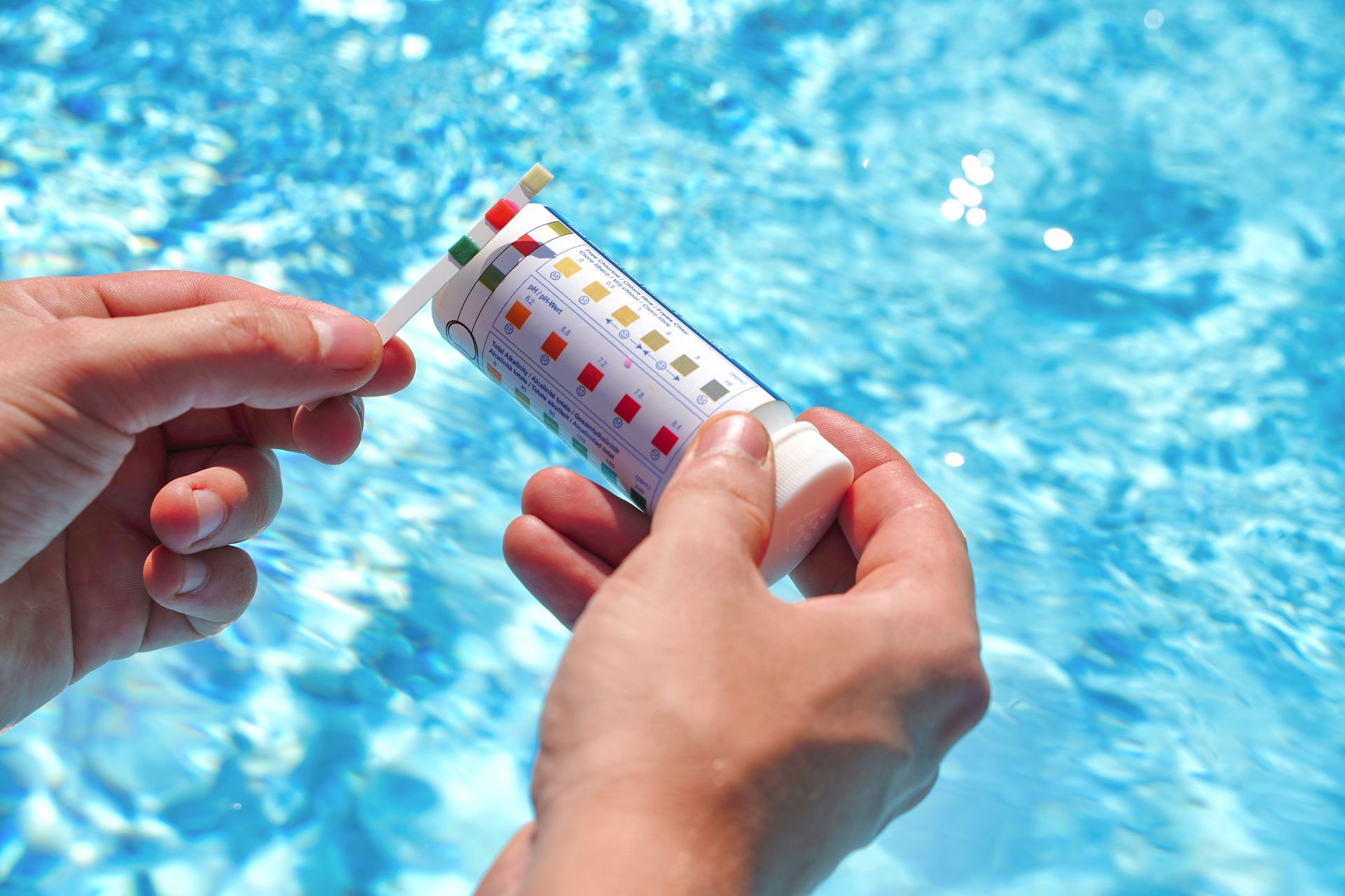 Hands hold a pool test strip against its color-coded comparison chart with a blue water background.