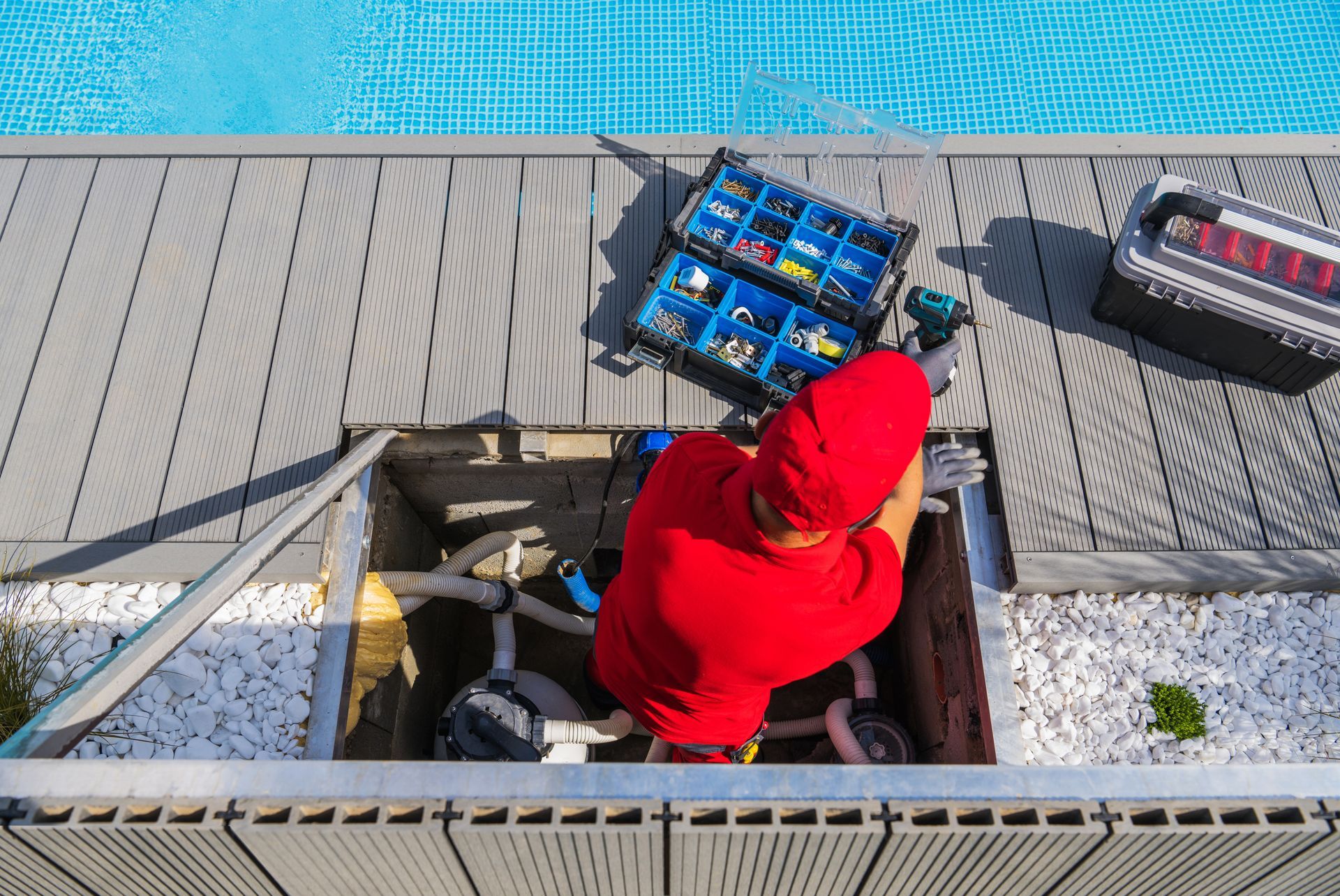 A worker in a red shirt and cap repairs pool equipment inside an open utility pit on a deck next to a swimming pool.