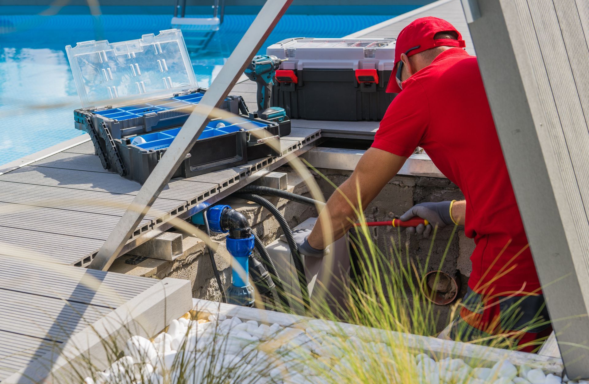A technician in a red uniform works on pool maintenance equipment in an open compartment beside a swimming pool.