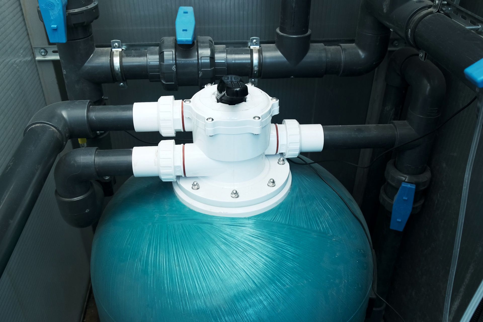 A blue pool sand filter tank with a white multiport valve connected by black plumbing pipes and blue shut-off valves.