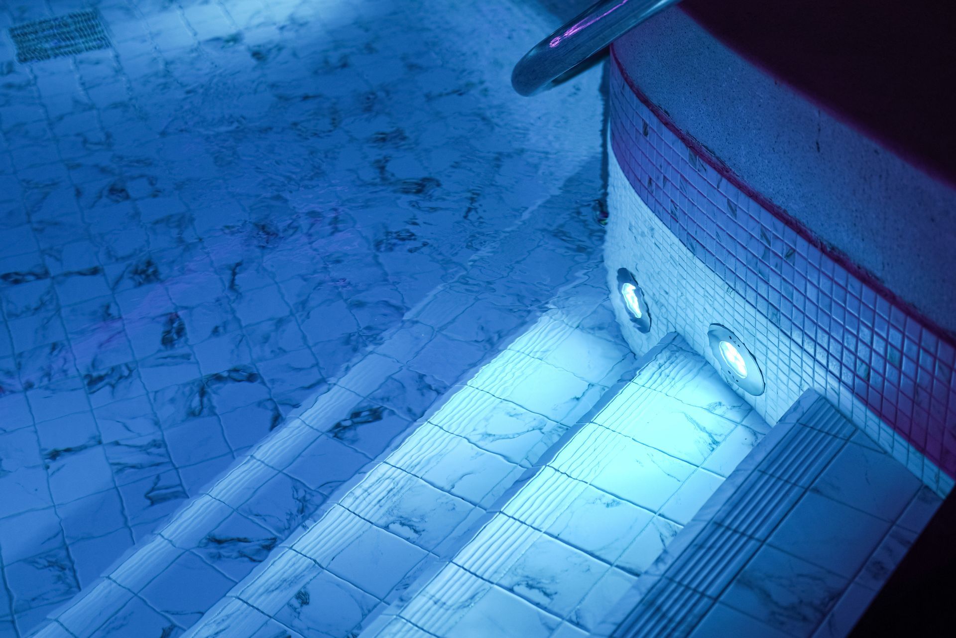 Underwater view of tiled swimming pool steps illuminated by bright blue lights.