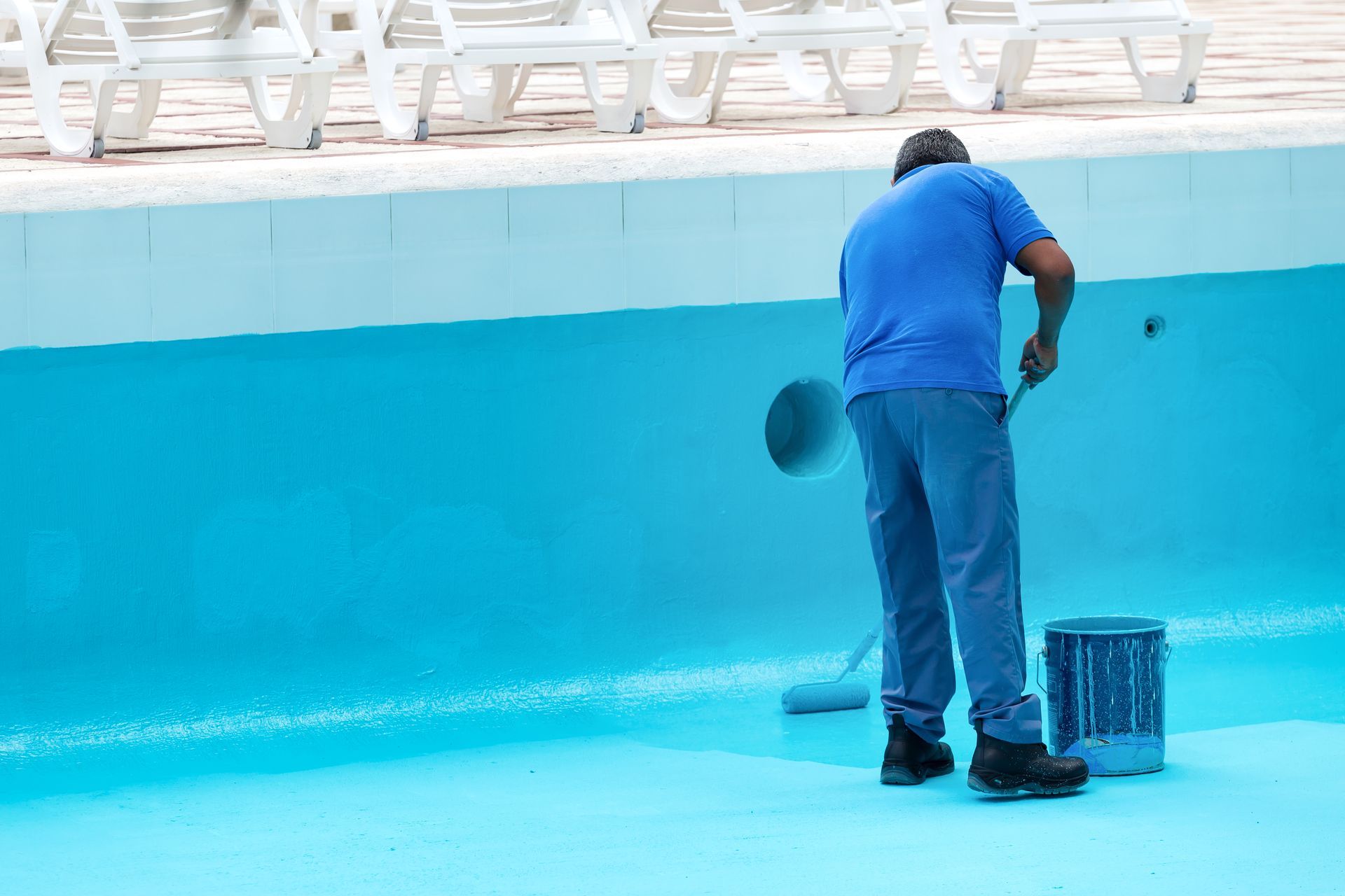 A person in blue clothes paints the floor of an empty swimming pool with a blue roller and a paint can.